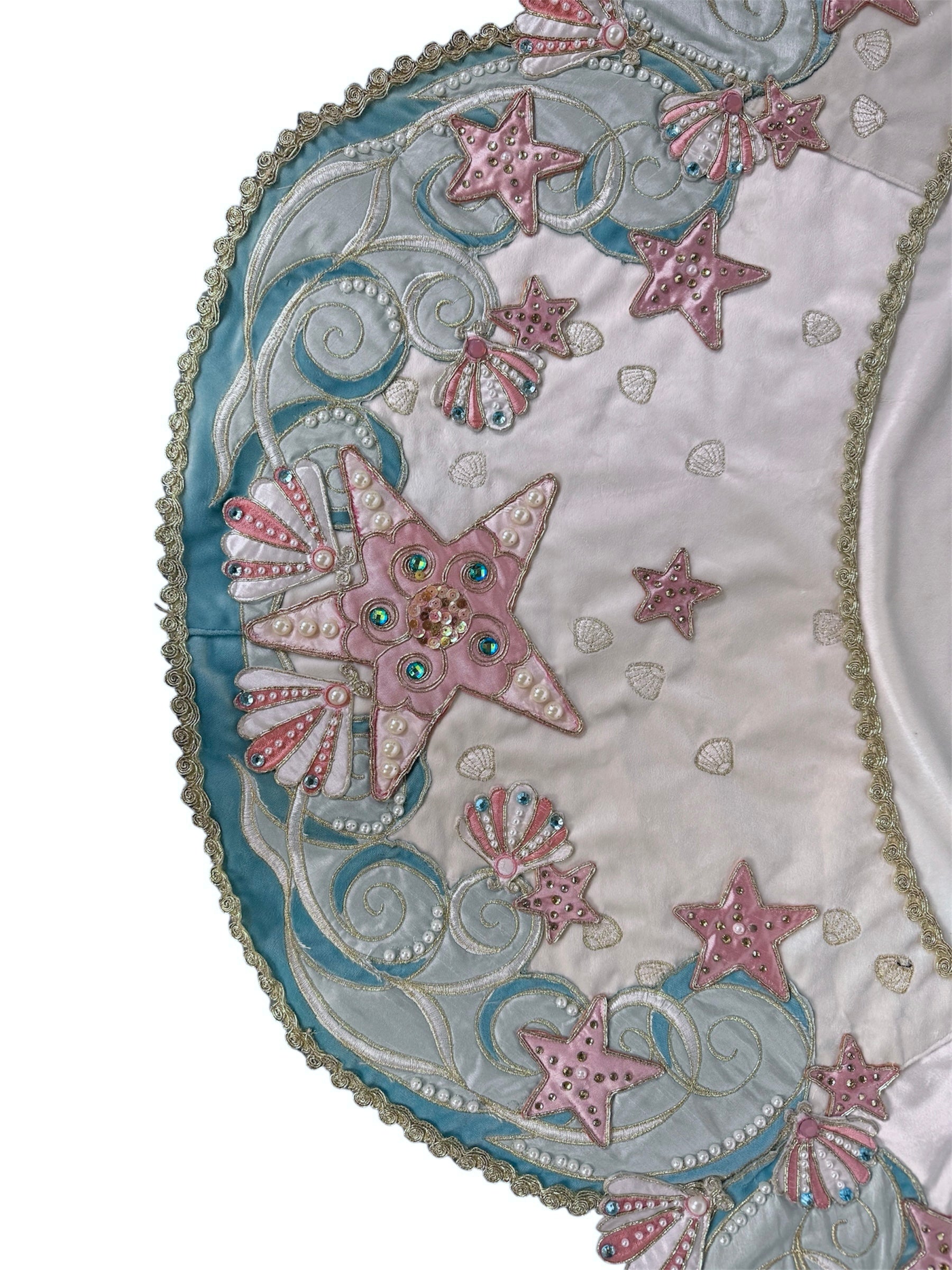 A round, cream-colored tablecloth featuring a dramatic, scalloped border shaped like six large flower petals. The border is intricately embroidered with a coastal motif including pink starfish, seashells, and teal-blue swirling waves. Gold beaded accents line the inner circular seam and the outer edges.
A long, narrow cream runner with matching scalloped, wavy edges. It is adorned with the same nautical embroidery of pink starfish and seashells scattered across the center and concentrated along the edge.