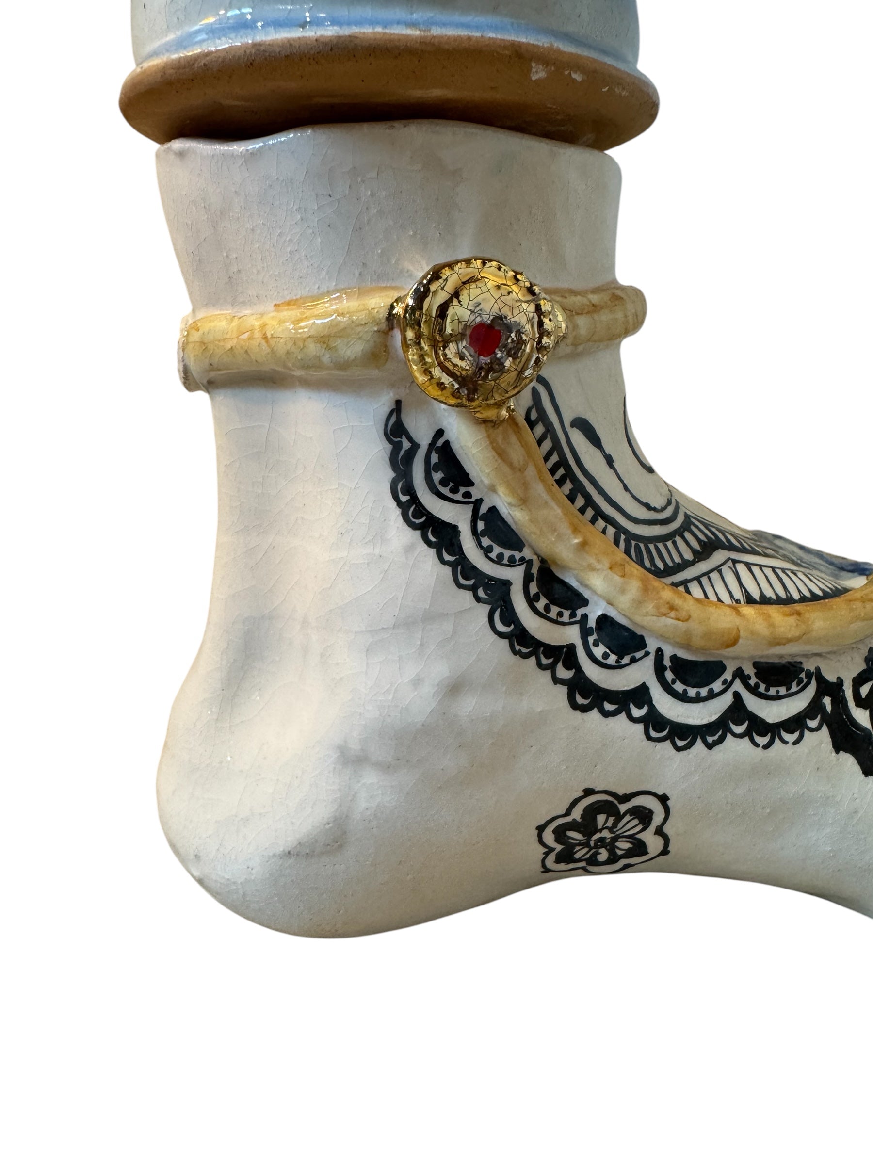 Handcrafted ceramic sculpture in the shape of a foot, decorated with intricate black and white patterns, 24KT gold accents, and topped with a blue lid featuring a gilt fish and head emerging from stylized waves.