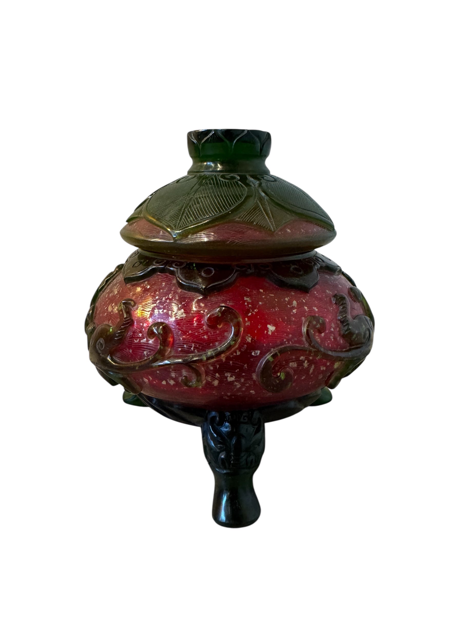 Close-up of a Qianlong-style Peking overlay glass censer. The red glass body is decorated with a carved green overlay design. The censer has a green lotus-carved lid and sits on three feet. A seal mark is visible on the base