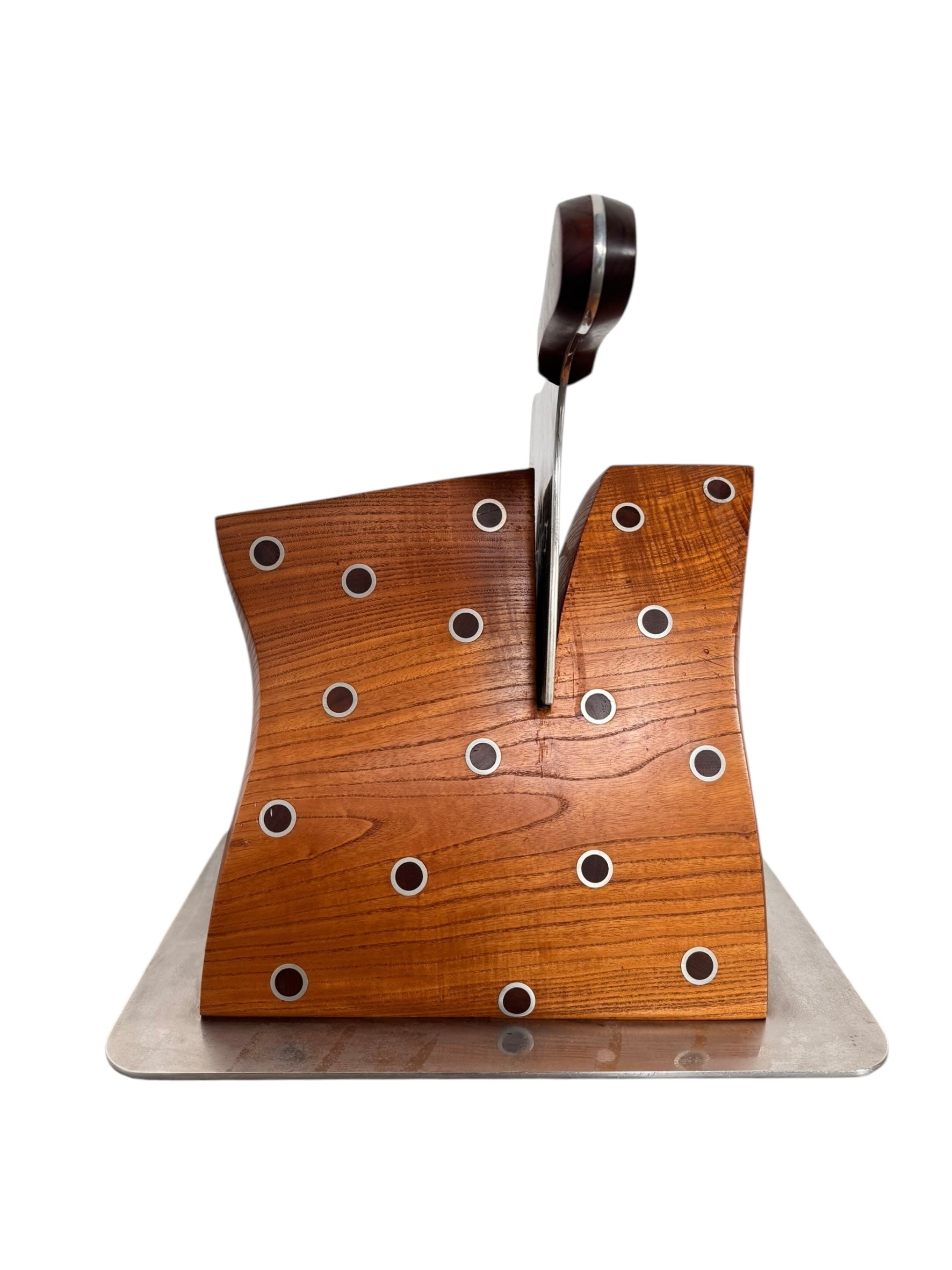 A wooden sculpture with a highly polished, reddish-brown grain, featuring two main asymmetrical, curved sections that converge at the top. Circular metal inlays with black centers are embedded across the surface of both wooden sections. A flat, polished metal blade-like element is inserted vertically between the two wooden sections, extending above the top surface. The sculpture rests on a metallic base.