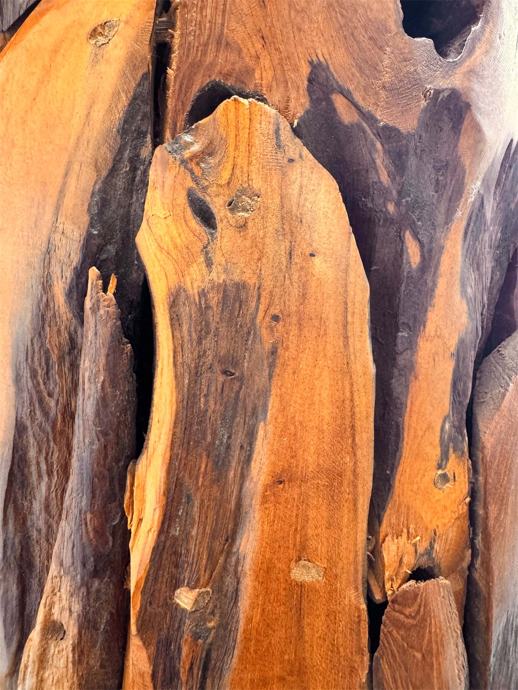 Two table lamps made from repurposed teak wood vases. The bases are tall and tapered, featuring a rugged, mosaic-like texture composed of various pieces of dark, polished teak wood with visible natural grain and crevices. Each lamp is topped with a large, warm-toned conical shade that is currently wrapped in protective clear plastic, emitting a soft, golden glow. The lamps are positioned on a clean white surface against a neutral background.
