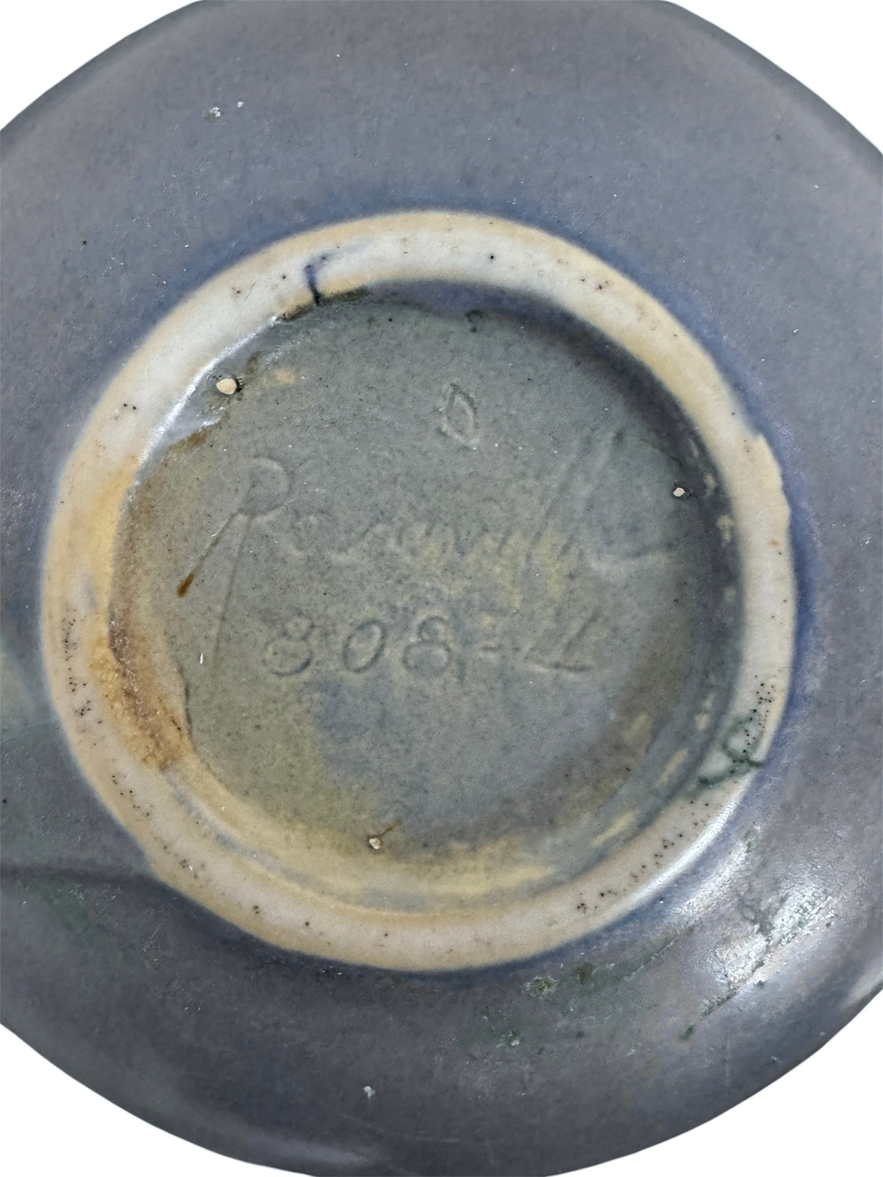 A squat, rounded vintage Roseville pottery vase in a light teal-blue matte glaze. The vase features two small, angular geometric handles on its shoulders and a flared rim. Embossed on the front is a cluster of textured, green and pale-yellow thornapple blossoms tucked against sharp, serrated green leaves. The surface shows a subtle, fine network of age-related crazing lines throughout the glaze.