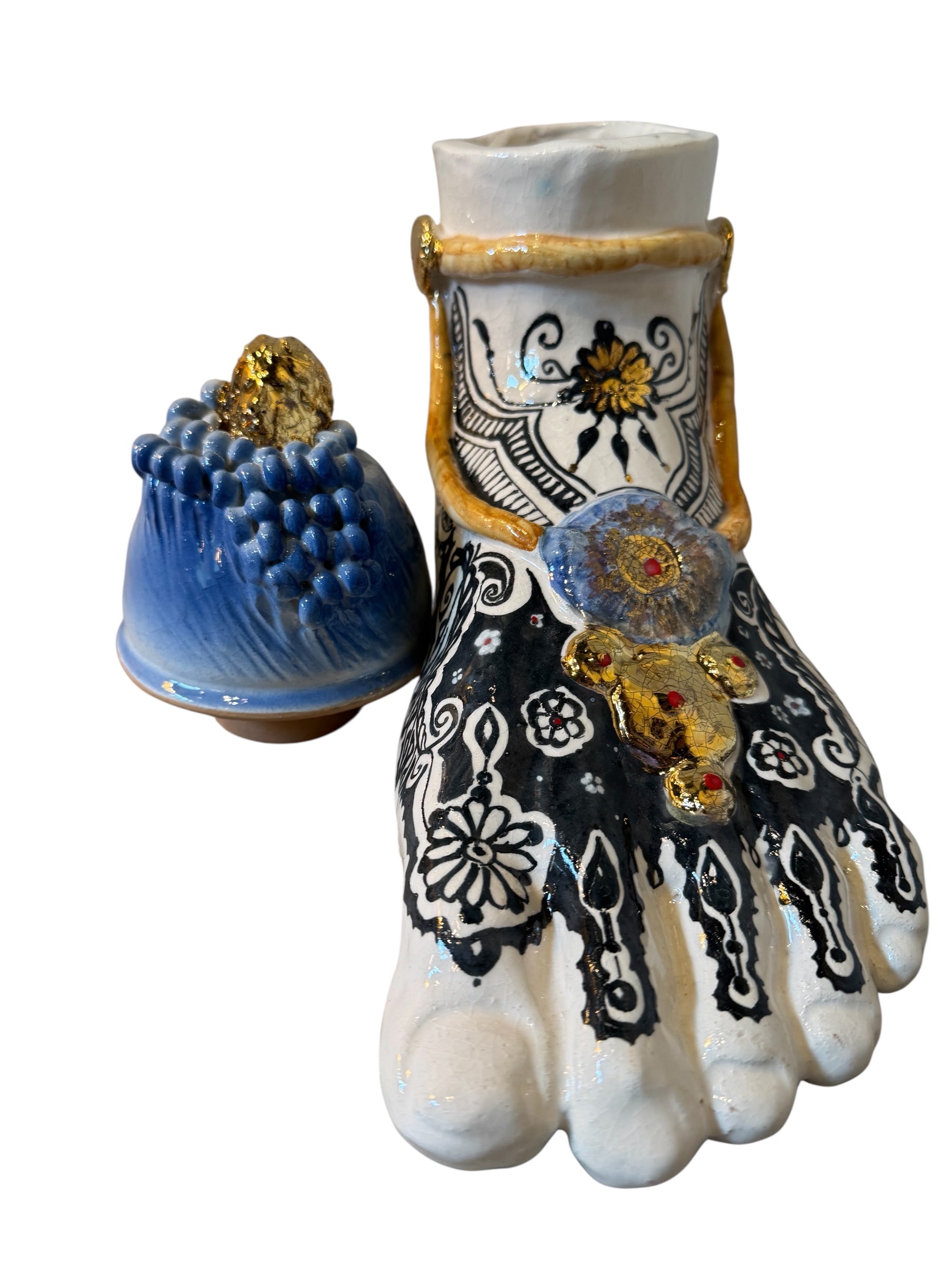 Handcrafted ceramic sculpture in the shape of a foot, decorated with intricate black and white patterns, 24KT gold accents, and topped with a blue lid featuring a gilt fish and head emerging from stylized waves.