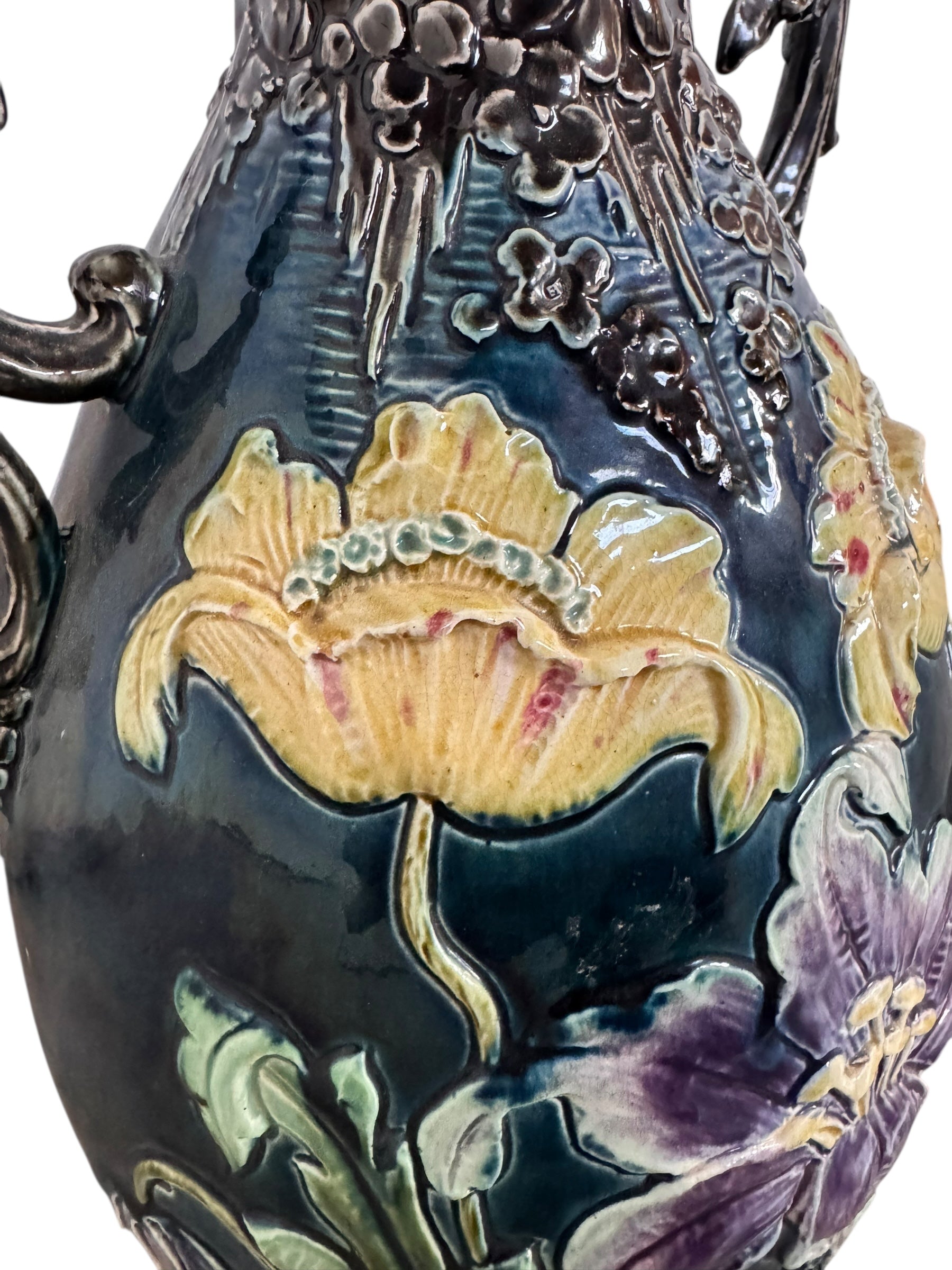 An ornate, dark glazed ceramic Art Nouveau vase with two handles and raised floral designs in yellow, purple, and green.