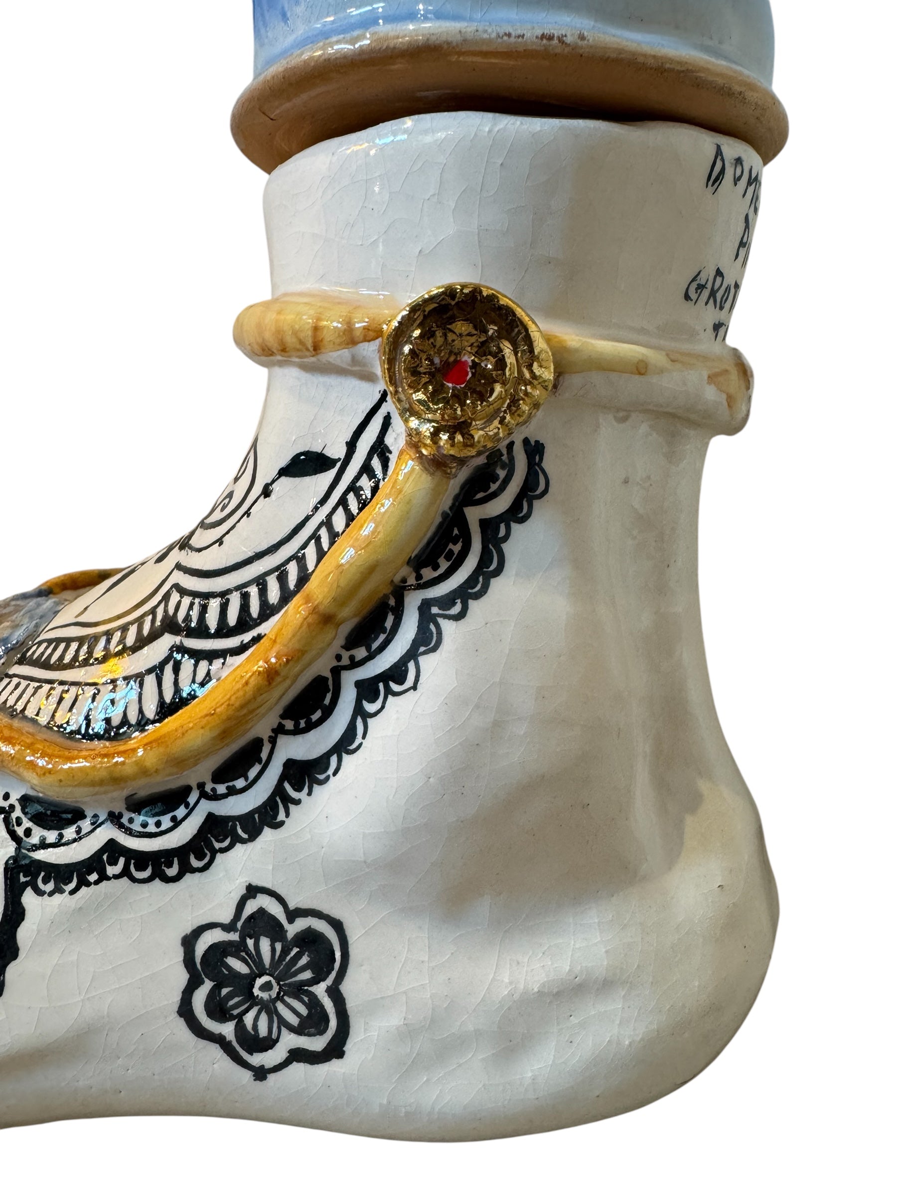 Handcrafted ceramic sculpture in the shape of a foot, decorated with intricate black and white patterns, 24KT gold accents, and topped with a blue lid featuring a gilt fish and head emerging from stylized waves.