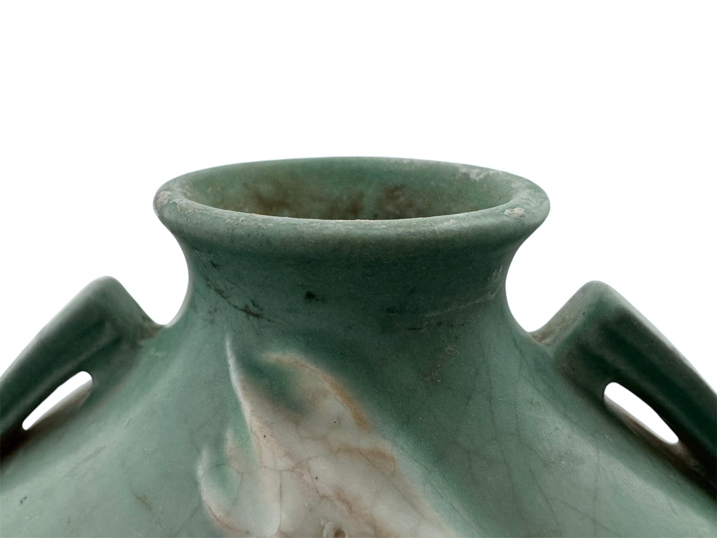 A squat, rounded vintage Roseville pottery vase in a light teal-blue matte glaze. The vase features two small, angular geometric handles on its shoulders and a flared rim. Embossed on the front is a cluster of textured, green and pale-yellow thornapple blossoms tucked against sharp, serrated green leaves. The surface shows a subtle, fine network of age-related crazing lines throughout the glaze.