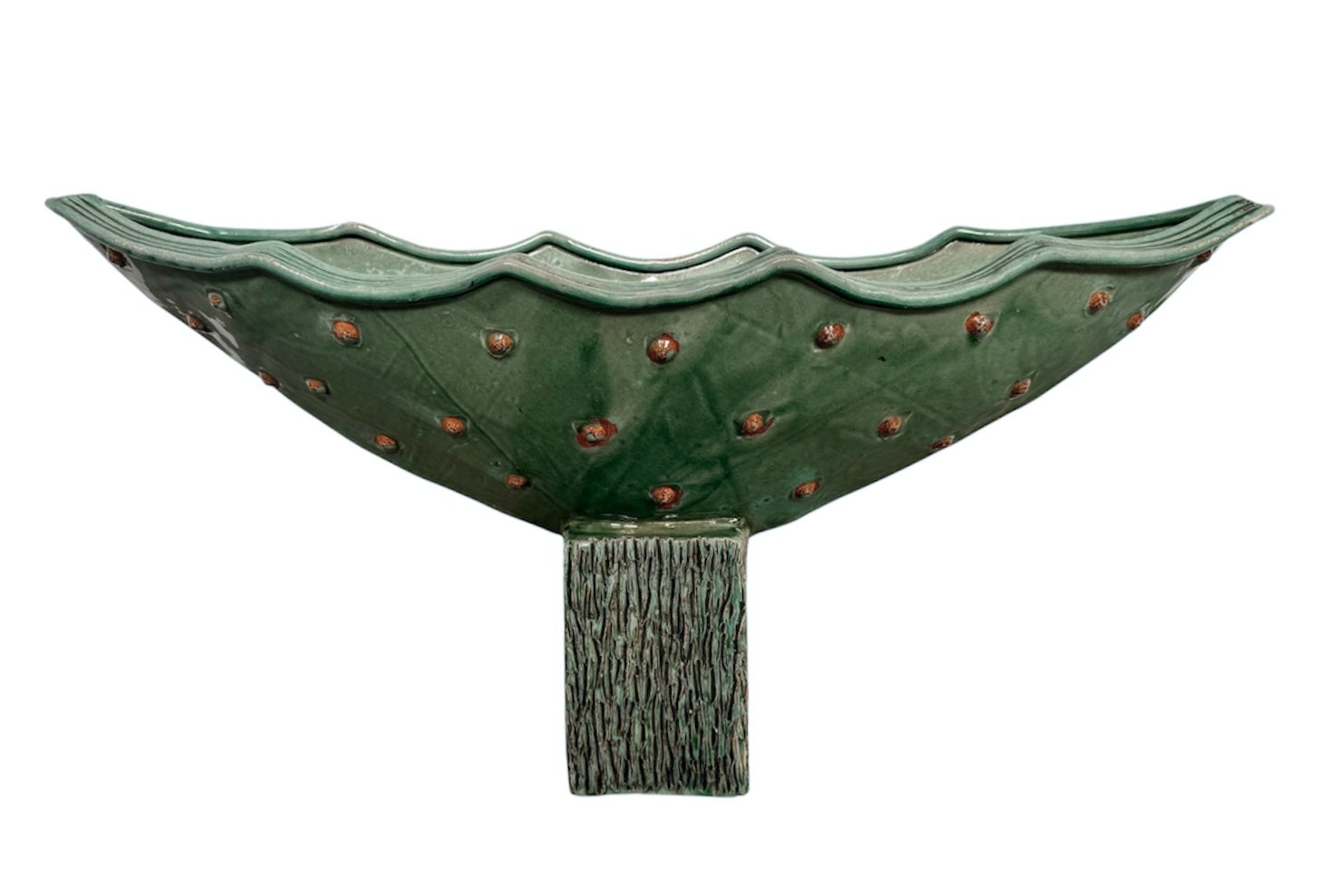 curved ceramic piece resembling a green pod, supported by a block plinth. The seafoam-green ceramic structure extends wide on either side, curving upward to form a wavy, boat-like shape. The outer surface is dotted with rows of small, raised, brown-orange, acorn-like protrusions. The top rim is layered with parallel, grooved ridges. The base is a vertical rectangular block with a heavily textured, bark-like surface in a dark green and black finish.