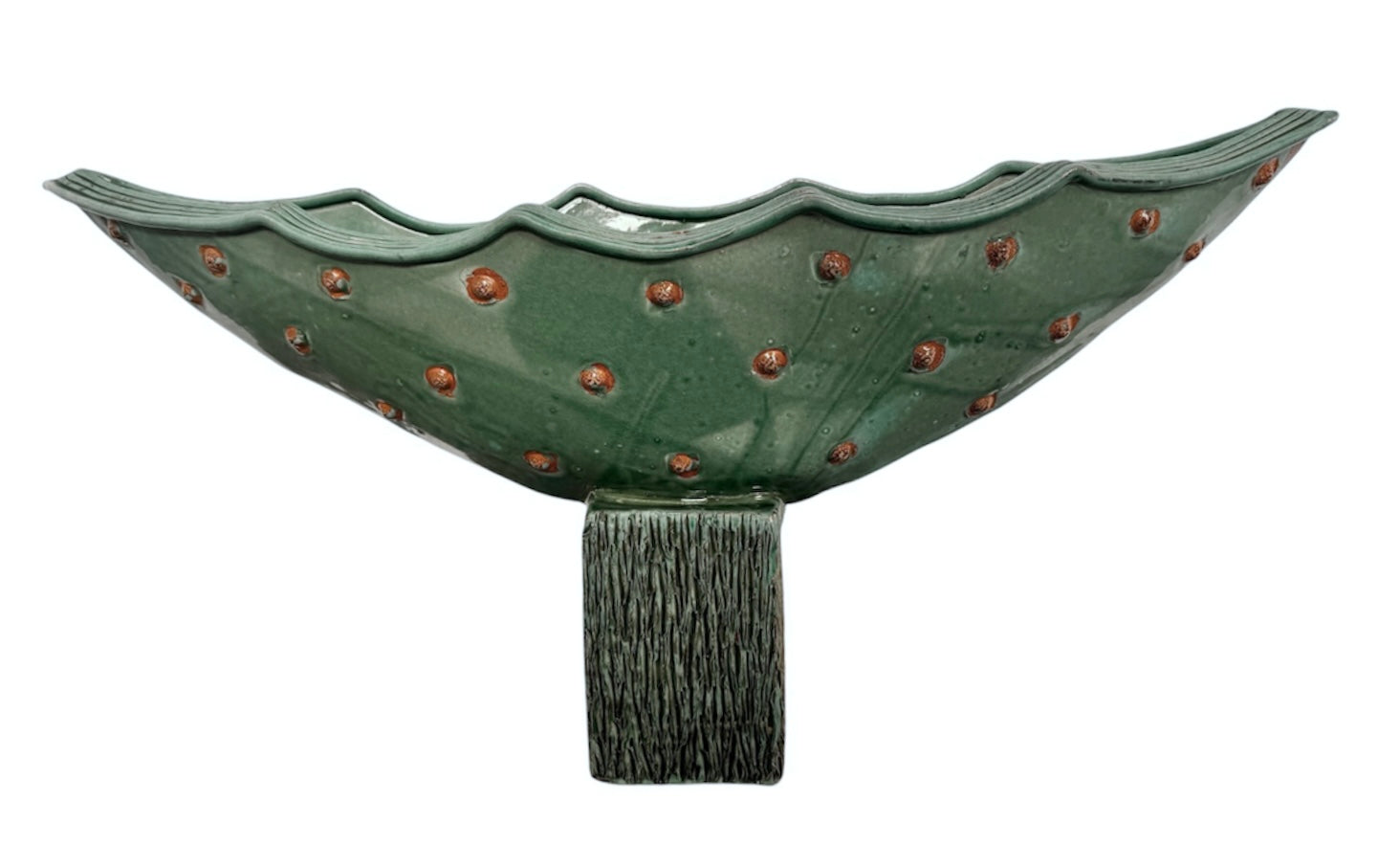 curved ceramic piece resembling a green pod, supported by a block plinth. The seafoam-green ceramic structure extends wide on either side, curving upward to form a wavy, boat-like shape. The outer surface is dotted with rows of small, raised, brown-orange, acorn-like protrusions. The top rim is layered with parallel, grooved ridges. The base is a vertical rectangular block with a heavily textured, bark-like surface in a dark green and black finish.