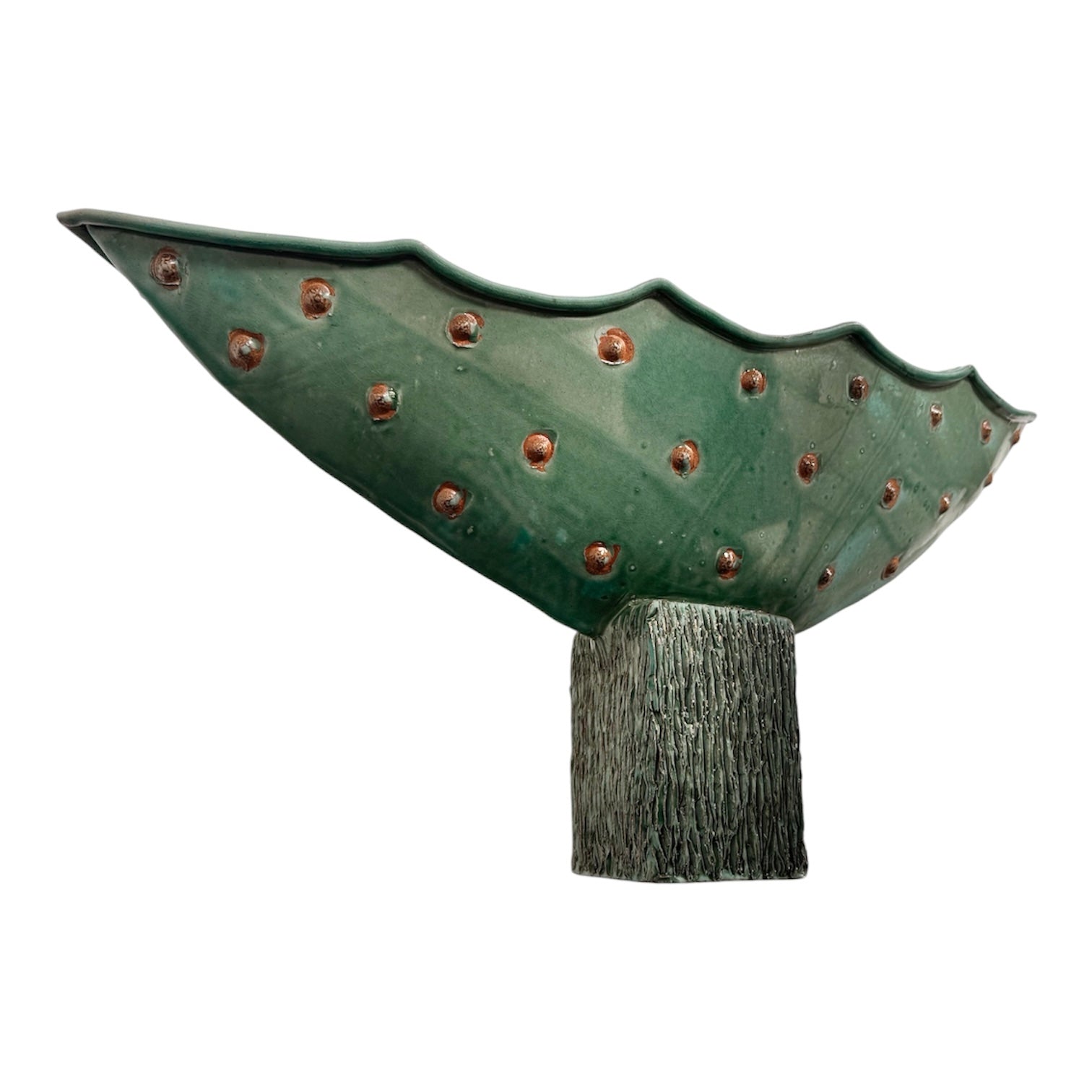 curved ceramic piece resembling a green pod, supported by a block plinth. The seafoam-green ceramic structure extends wide on either side, curving upward to form a wavy, boat-like shape. The outer surface is dotted with rows of small, raised, brown-orange, acorn-like protrusions. The top rim is layered with parallel, grooved ridges. The base is a vertical rectangular block with a heavily textured, bark-like surface in a dark green and black finish.
