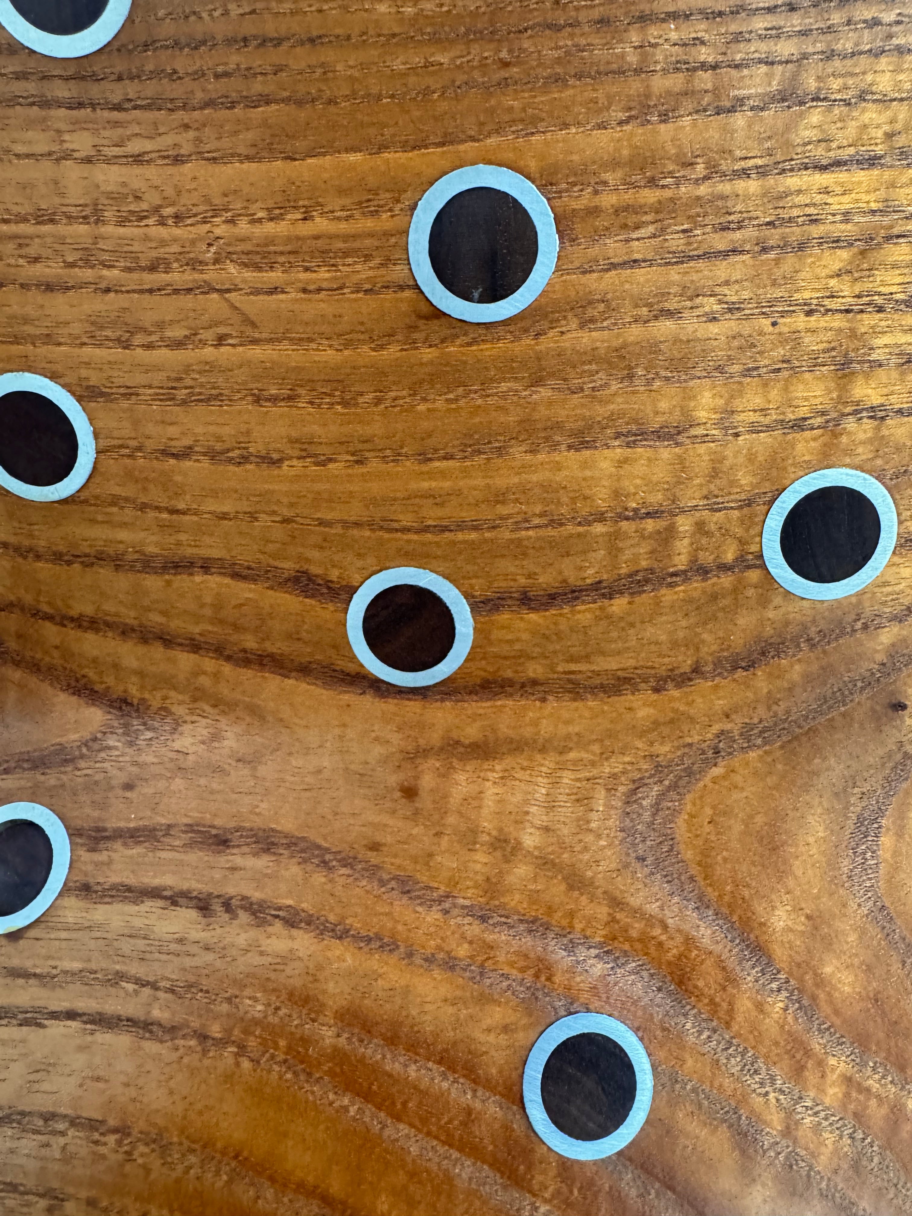 A wooden sculpture with a highly polished, reddish-brown grain, featuring two main asymmetrical, curved sections that converge at the top. Circular metal inlays with black centers are embedded across the surface of both wooden sections. A flat, polished metal blade-like element is inserted vertically between the two wooden sections, extending above the top surface. The sculpture rests on a metallic base.