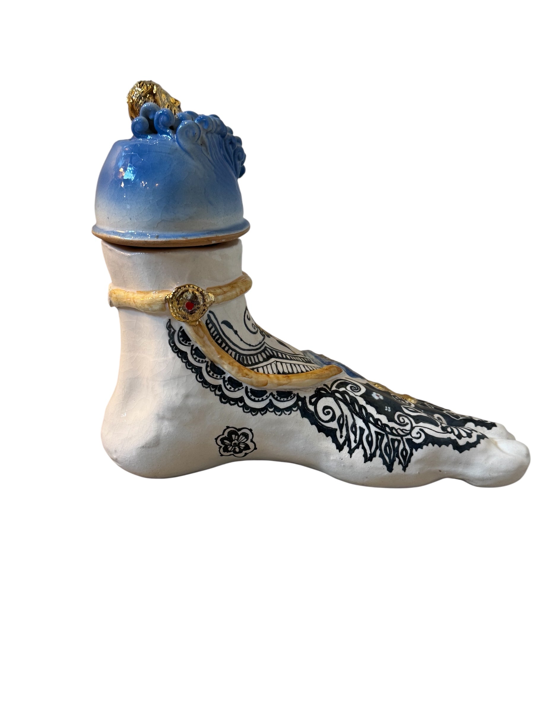 Handcrafted ceramic sculpture in the shape of a foot, decorated with intricate black and white patterns, 24KT gold accents, and topped with a blue lid featuring a gilt fish and head emerging from stylized waves.