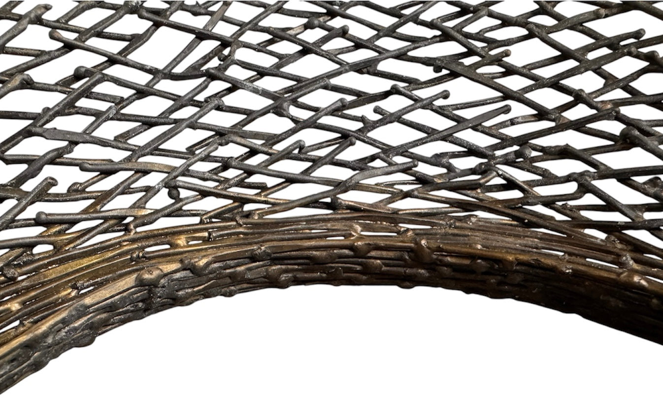 A round metal wall sculpture by Michael Aram, viewed from the front against a stark white background. The piece is composed of a dense, intricate lattice of fine, dark metal wire or twigs woven together to create a textured, web-like pattern. The structure curves inward like a funnel or a vortex, sloping down toward a clean, circular opening in the exact center, creating a strong sense of depth and dimensionality.