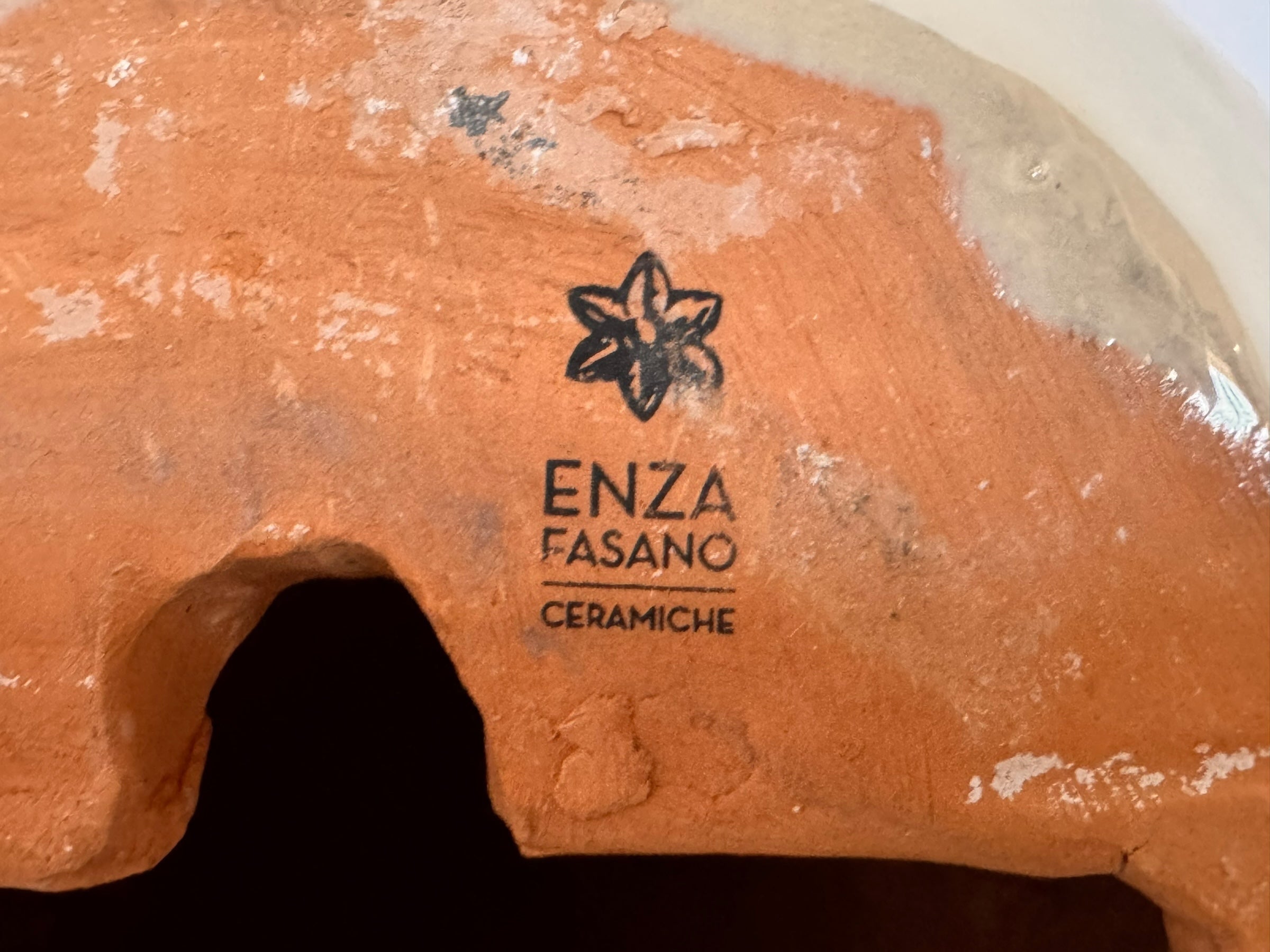 A ceramic sculpture of a bulldog's head by Enza Fasano. The piece features a glossy, glazed finish with a sophisticated color palette of muted greys, creams, and earthy brown tones arranged in bold, abstract striped patterns across the face and ears. The bulldog has characteristic folded ears, a prominent snout, and a soulful, slightly downturned expression. The sculpture is set against a clean, white background, highlighting its smooth texture and reflective surface.