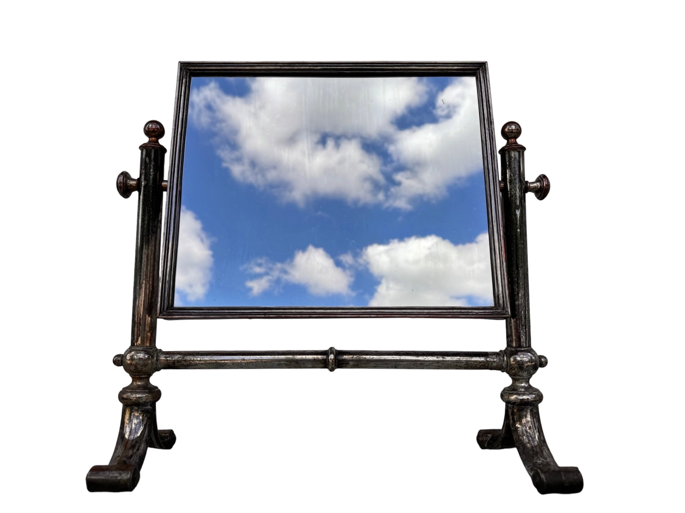 The antique Chevalier Mirror features a rectangular swing frame supported by two turned posts on decorative trestle feet. The mirror stands on four scrolled feet and features its original tarnished, silvered finish. The front of the mirror reflects a bright blue sky with wispy white clouds. The original dark brown backing is still attached to the frame.