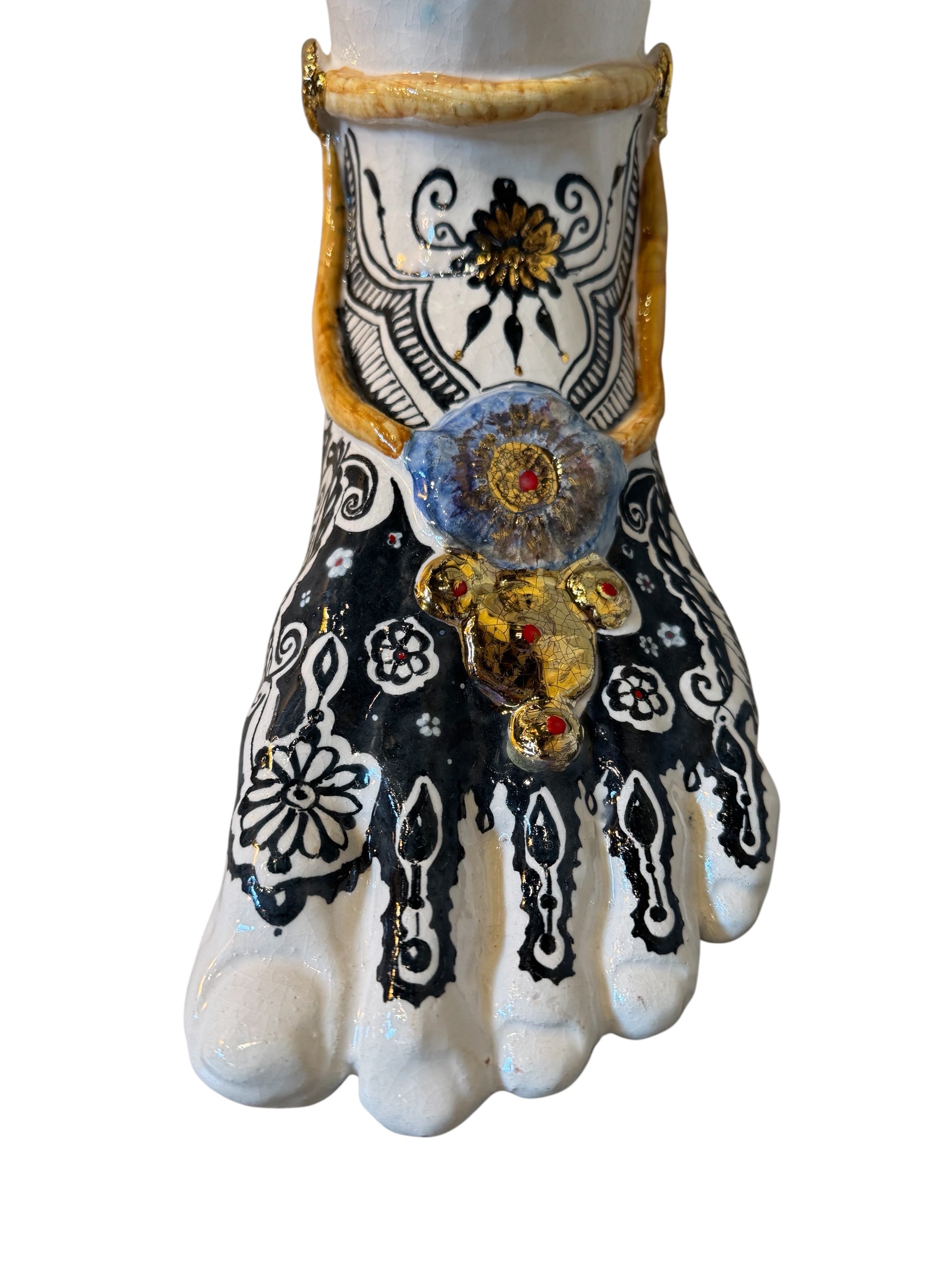 Handcrafted ceramic sculpture in the shape of a foot, decorated with intricate black and white patterns, 24KT gold accents, and topped with a blue lid featuring a gilt fish and head emerging from stylized waves.