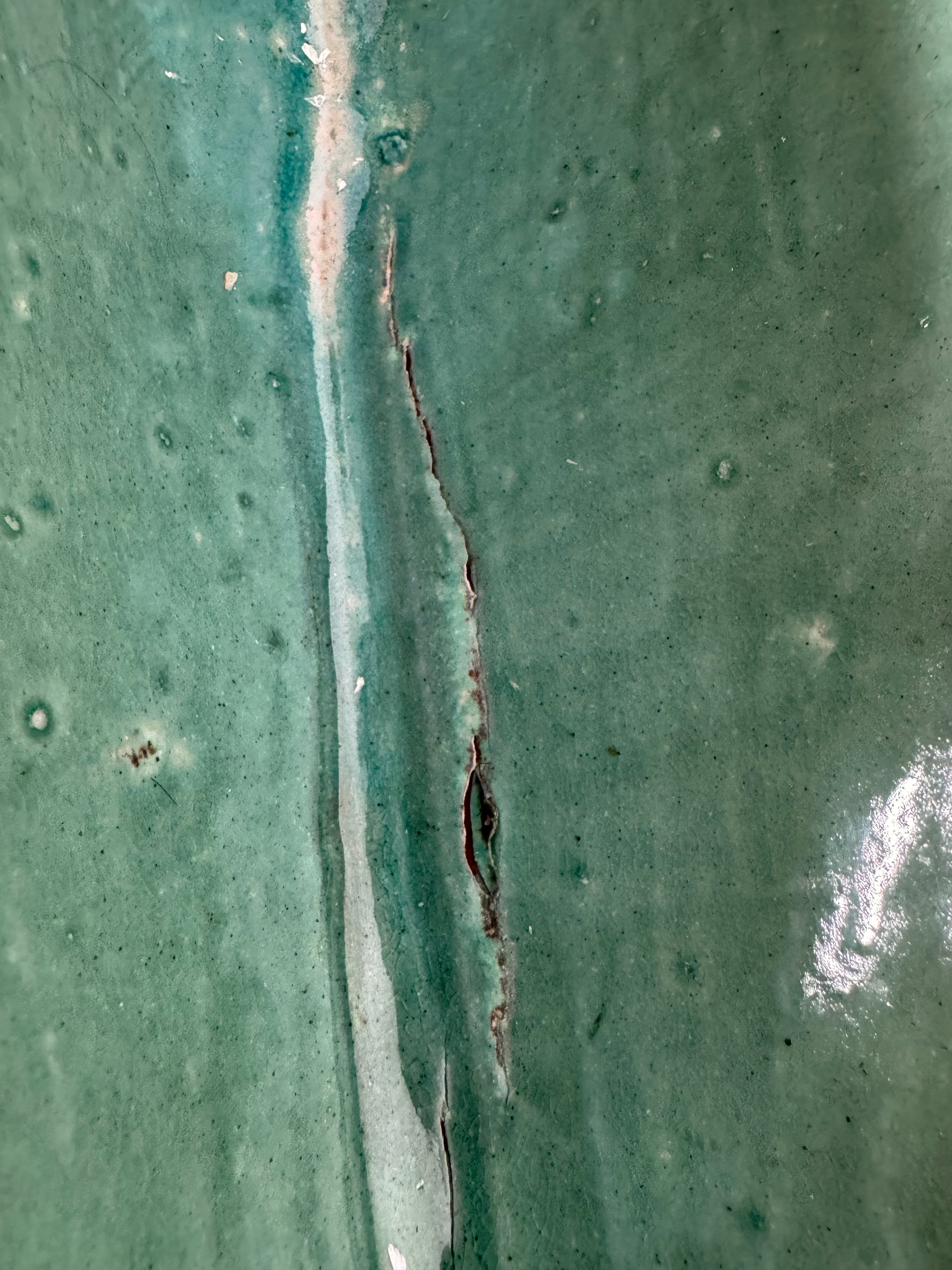 curved ceramic piece resembling a green pod, supported by a block plinth. The seafoam-green ceramic structure extends wide on either side, curving upward to form a wavy, boat-like shape. The outer surface is dotted with rows of small, raised, brown-orange, acorn-like protrusions. The top rim is layered with parallel, grooved ridges. The base is a vertical rectangular block with a heavily textured, bark-like surface in a dark green and black finish.
