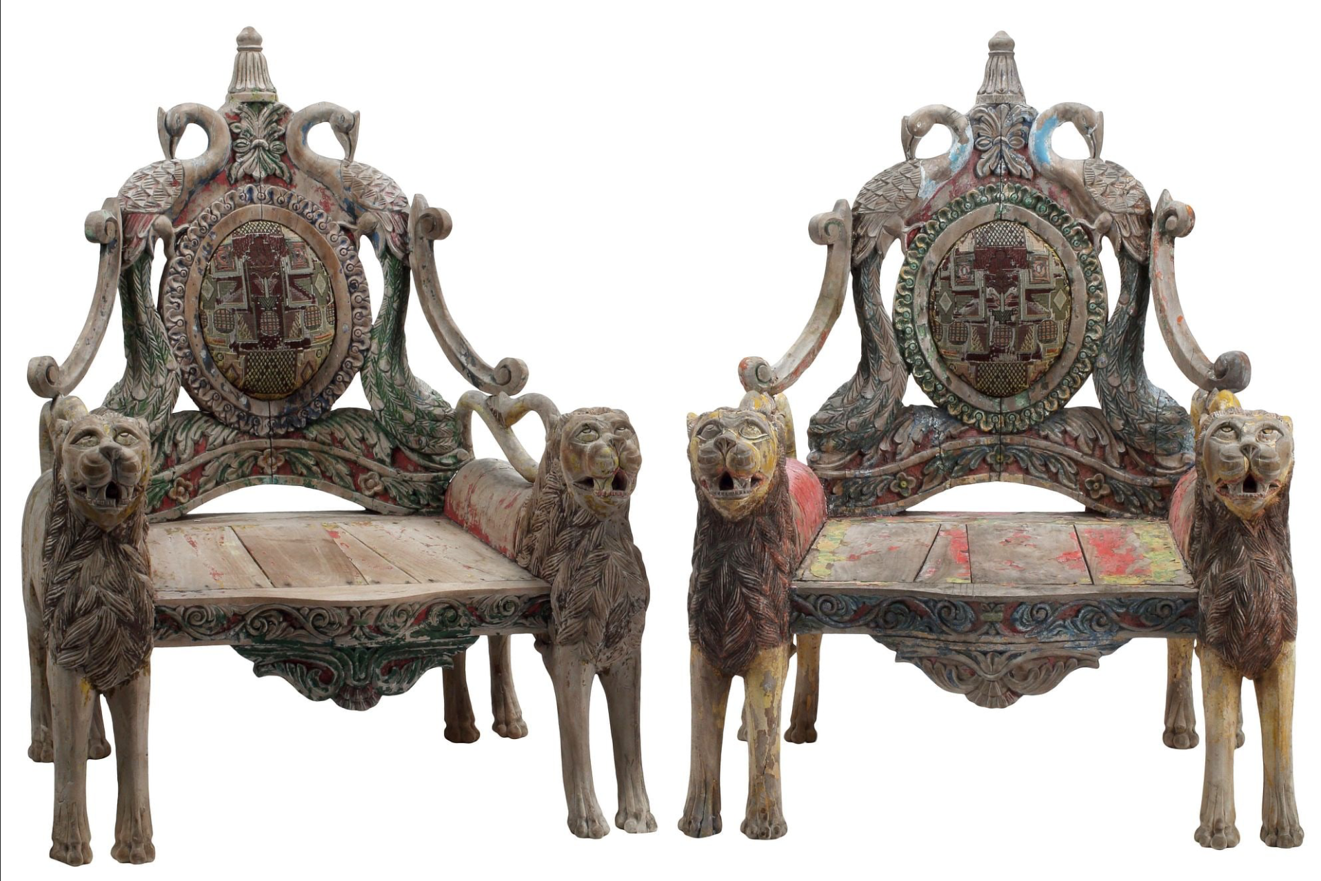A pair of antique, heavily carved Anglo-Indian style throne chairs featuring lion figures as legs and arm supports, and peacock motifs on the backrests. The chairs have a distressed, multi-colored painted finish.