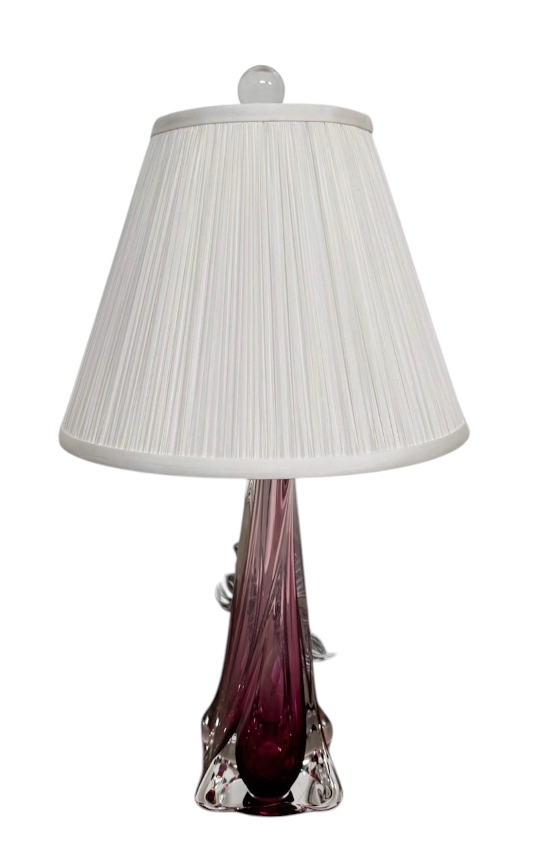 A tall, Mid-Century Modern sculptural table lamp featuring a hand-blown art glass base. The base is crafted in the Sommerso style, with a deep amethyst purple core encased in a thick, clear crystal outer layer that twists upward in an organic, fluid motion. The lamp is topped with a white, knife-pleated empire shade and a clear glass ball finial. The overall design is elegant and slender, emphasizing the contrast between the saturated purple glass and the transparent, light-refracting crystal.