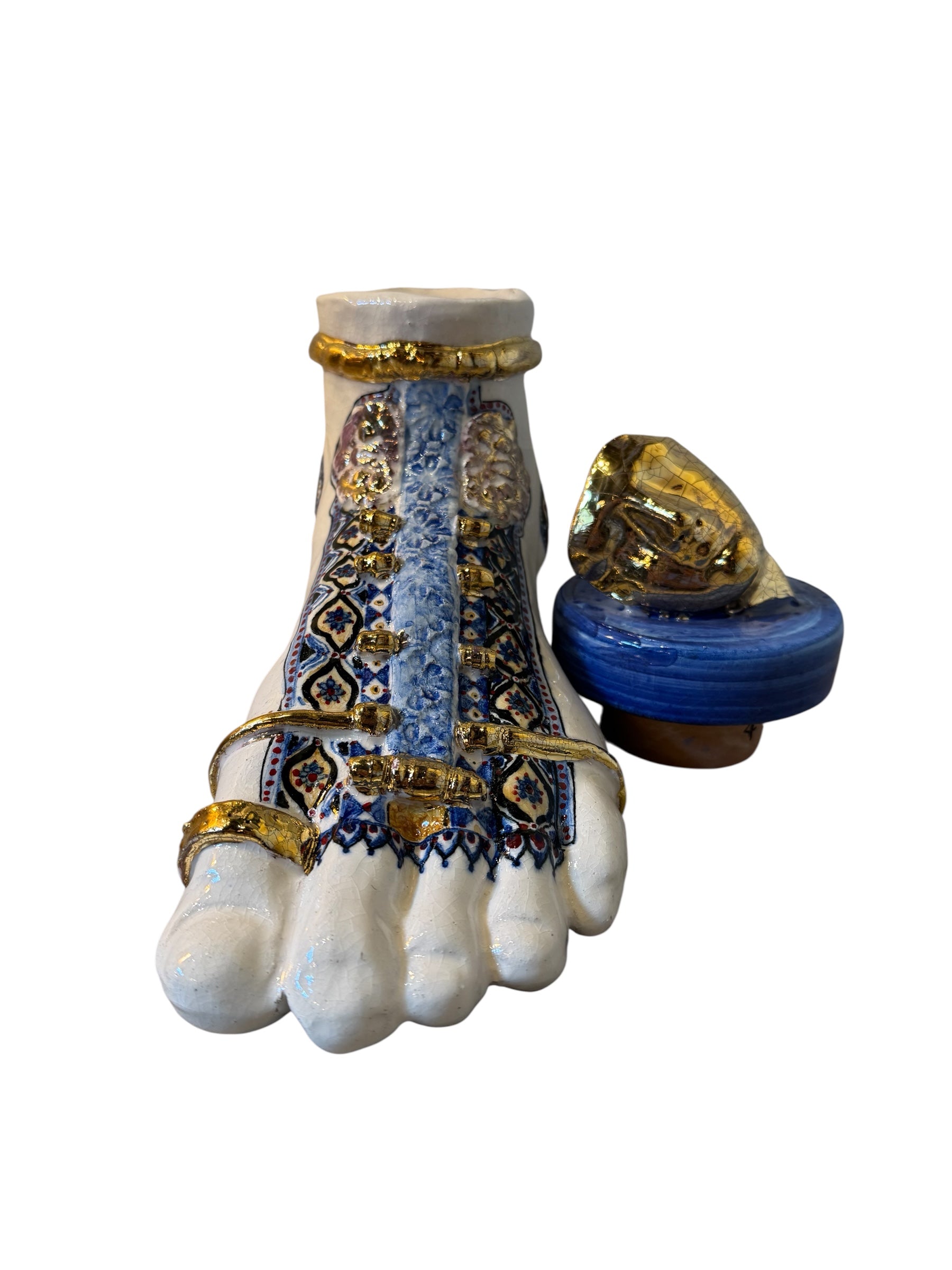 Close-up of a ceramic foot sculpture by Domenico Pinto. The foot is decorated with intricate hand-painted designs inspired by Apulian folklore and accented with 24KT gold. A stylized gilt mask sits atop the foot.