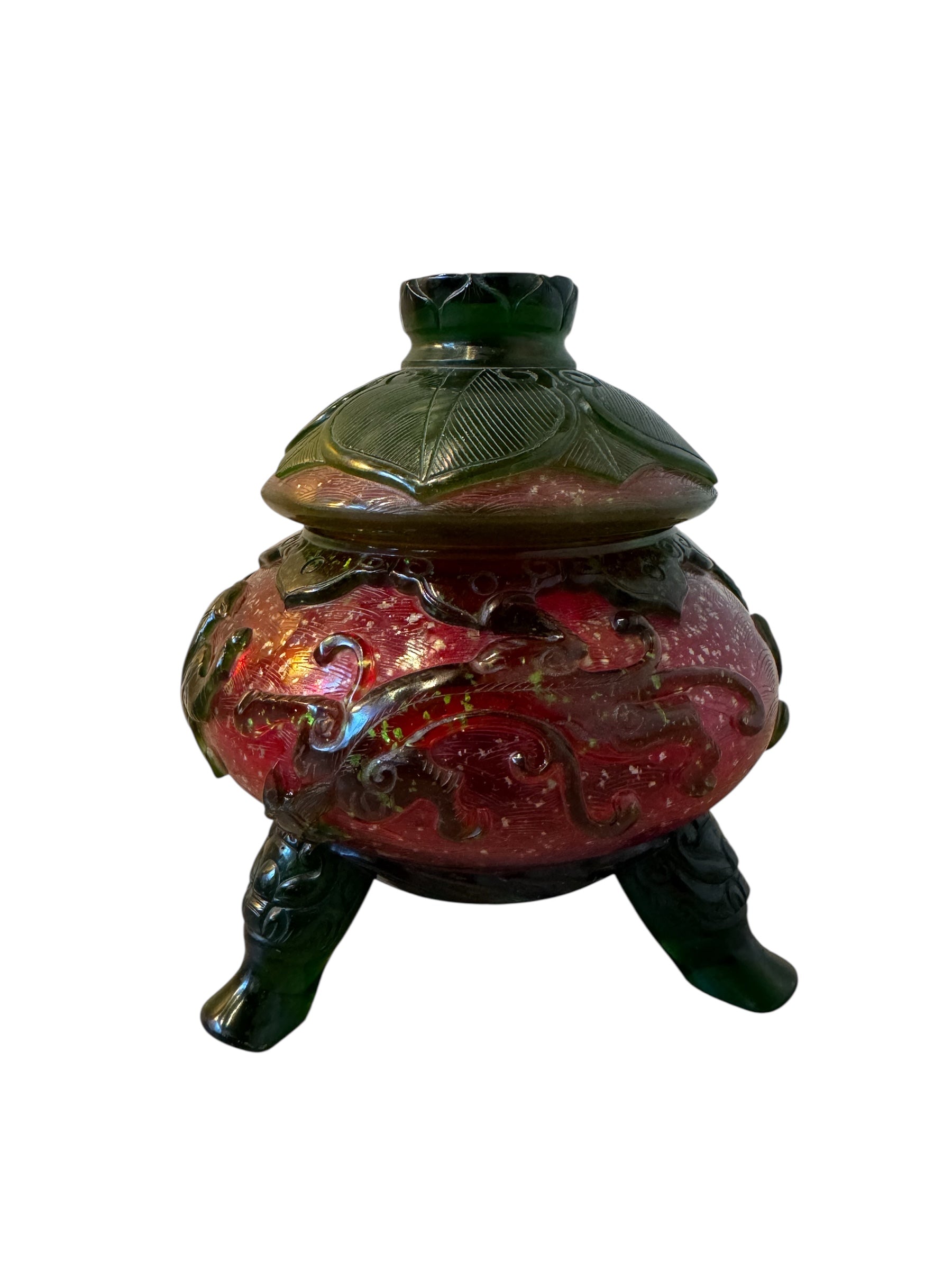 Close-up of a Qianlong-style Peking overlay glass censer. The red glass body is decorated with a carved green overlay design. The censer has a green lotus-carved lid and sits on three feet. A seal mark is visible on the base