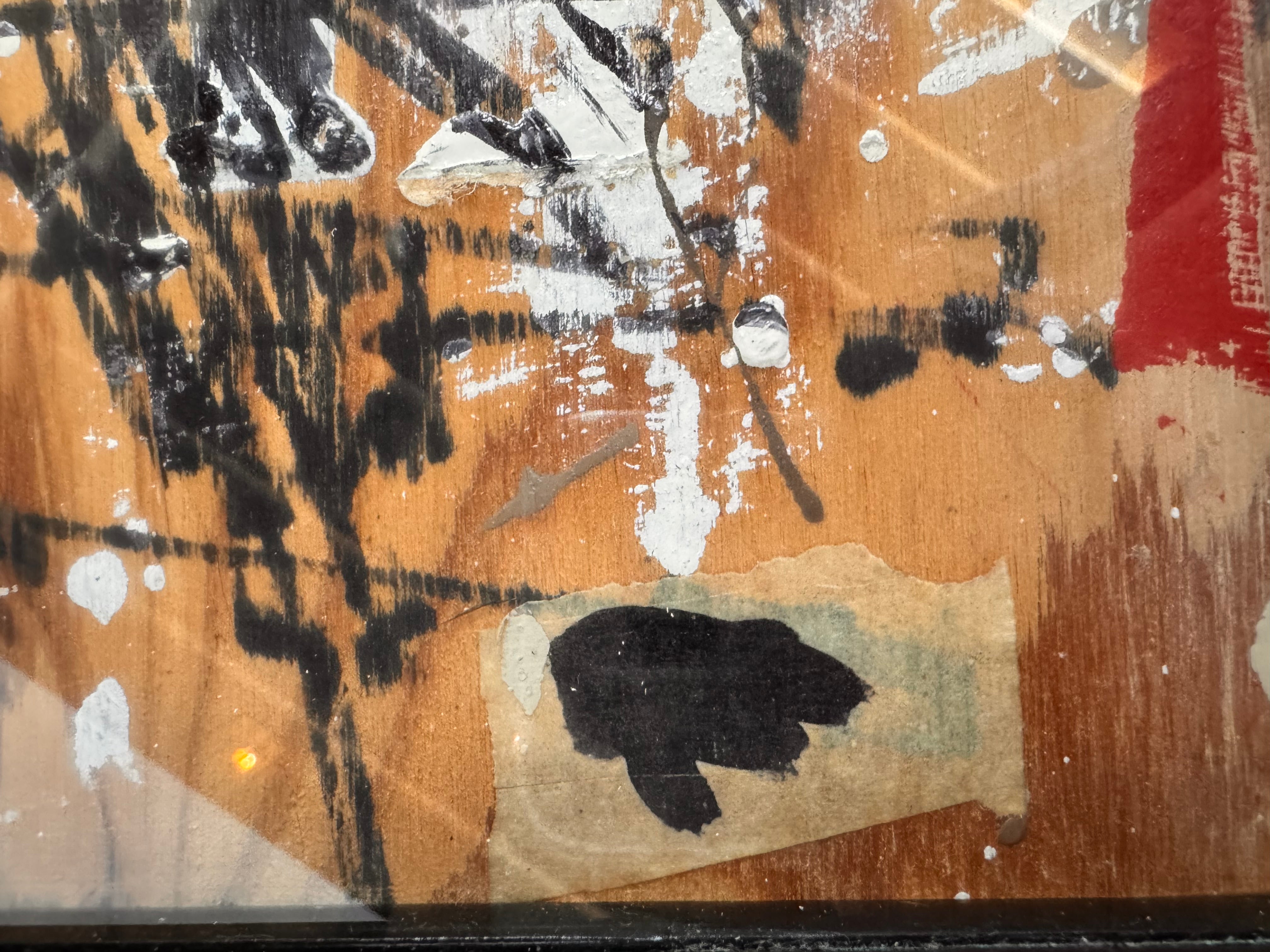 horizontal rectangular mixed media abstract artwork by J. Rogers, dated 2010, presented in a thin black frame. The composition is set against a light-toned wood grain background and features a dynamic, gestural arrangement of thick white impasto, splatters of red and black paint, and aggressive black scribbles. A vertical strip of textured material is collaged near the center, while various washes of tan and cream create a sense of weathered depth across the wooden surface.