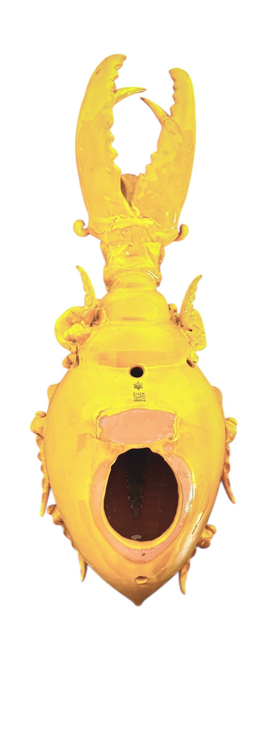 a large, high-gloss ceramic sculpture by Enza Fasano, shaped like a stylized beetle. The entire piece is finished in a warm, honey-yellow glaze. The beetle features prominent, curved mandibles at the front, a textured "collar" area, and a rounded, shell-like body with decorative ridges along the edges. The surface is smooth and reflective, catching the light across its organic contours.