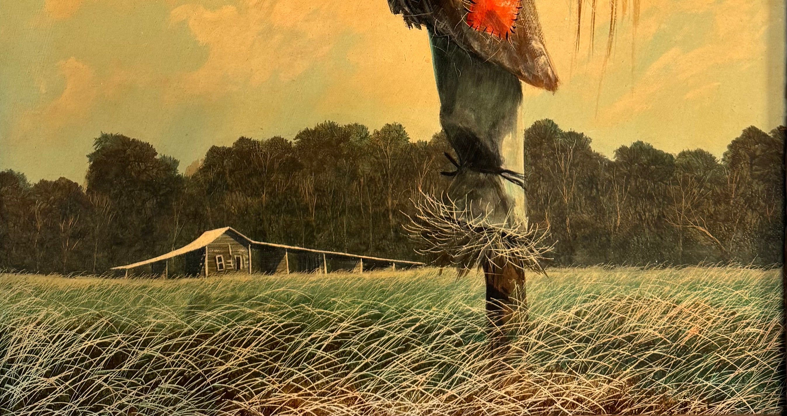 An oil painting on a wooden panel depicts a solitary scarecrow standing in a vast, grassy field under a pale yellow and blue sky. The scarecrow is shown from a three-quarters back view, wearing a brimmed hat and a tattered, patched brown coat with frayed straw bursting from its sleeves and edges. In the far left background, a long, white rustic barn sits before a dense line of green trees. The entire piece is enclosed in a thick, weathered, natural wood frame.