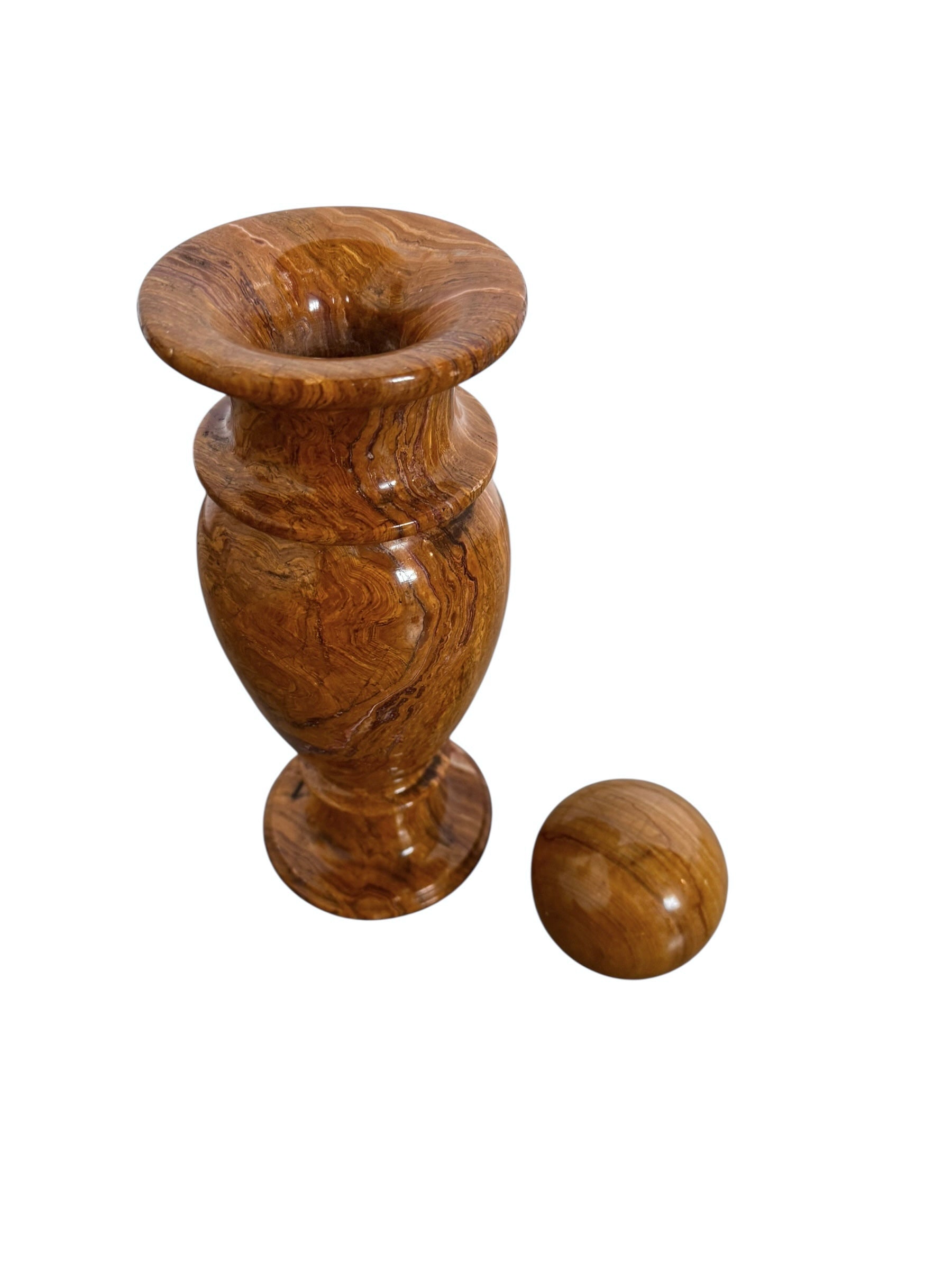 An elegant, hand-carved decorative vase made of polished Pakistani brown onyx. The vase features a classic, rounded urn shape with a tiered, flared neck and a solid matching spherical stone stopper resting on top. The stone displays rich, natural horizontal banding and swirling patterns in shades of honey, deep amber, rust-red, and cream. It stands on a small, integrated pedestal base against a clean white background, reflecting a high-gloss, mirror-like finish.