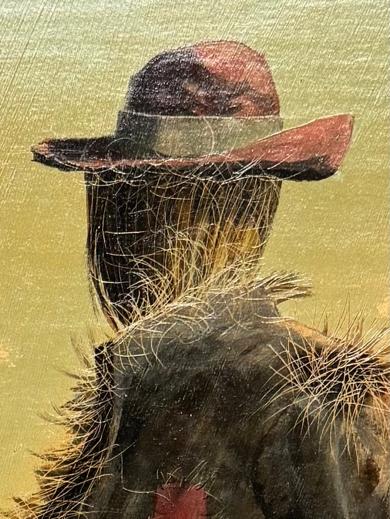 An oil painting on a wooden panel depicts a solitary scarecrow standing in a vast, grassy field under a pale yellow and blue sky. The scarecrow is shown from a three-quarters back view, wearing a brimmed hat and a tattered, patched brown coat with frayed straw bursting from its sleeves and edges. In the far left background, a long, white rustic barn sits before a dense line of green trees. The entire piece is enclosed in a thick, weathered, natural wood frame.