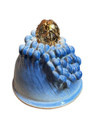 Handcrafted ceramic sculpture in the shape of a foot, decorated with intricate black and white patterns, 24KT gold accents, and topped with a blue lid featuring a gilt fish and head emerging from stylized waves.
