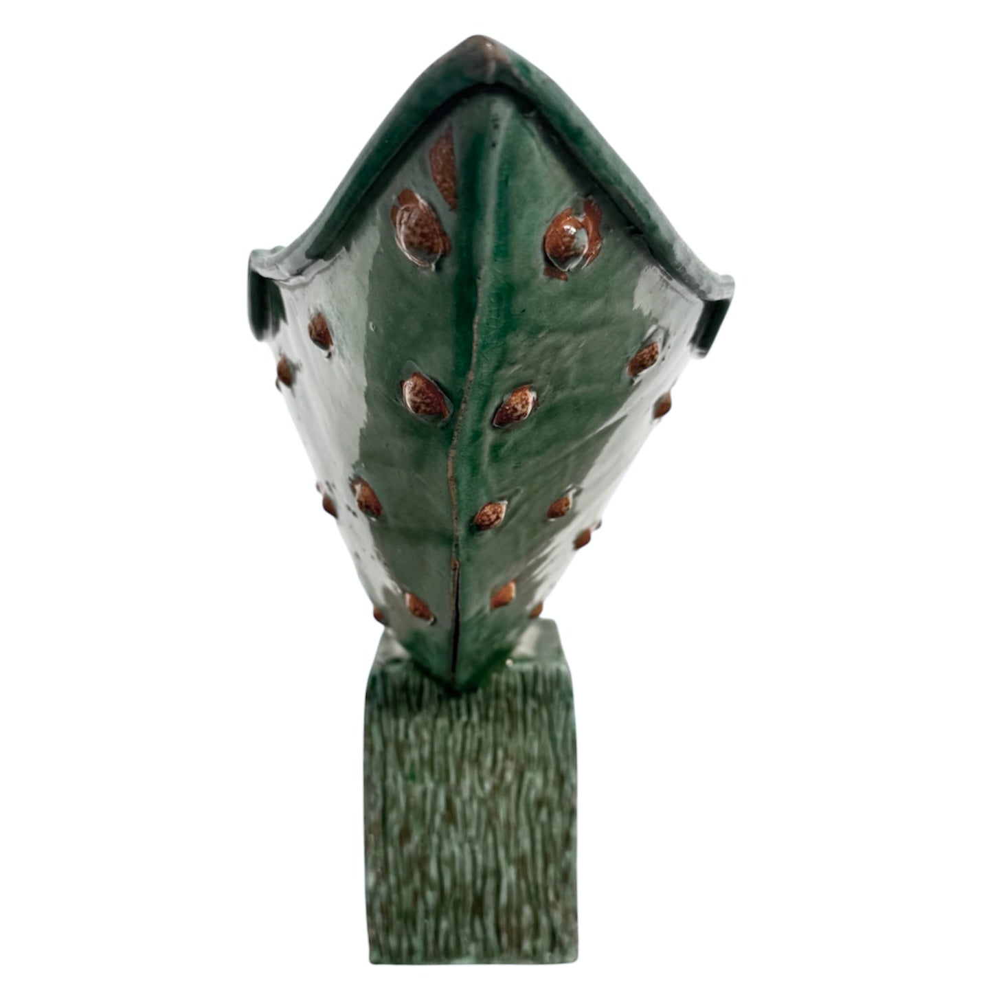 curved ceramic piece resembling a green pod, supported by a block plinth. The seafoam-green ceramic structure extends wide on either side, curving upward to form a wavy, boat-like shape. The outer surface is dotted with rows of small, raised, brown-orange, acorn-like protrusions. The top rim is layered with parallel, grooved ridges. The base is a vertical rectangular block with a heavily textured, bark-like surface in a dark green and black finish.