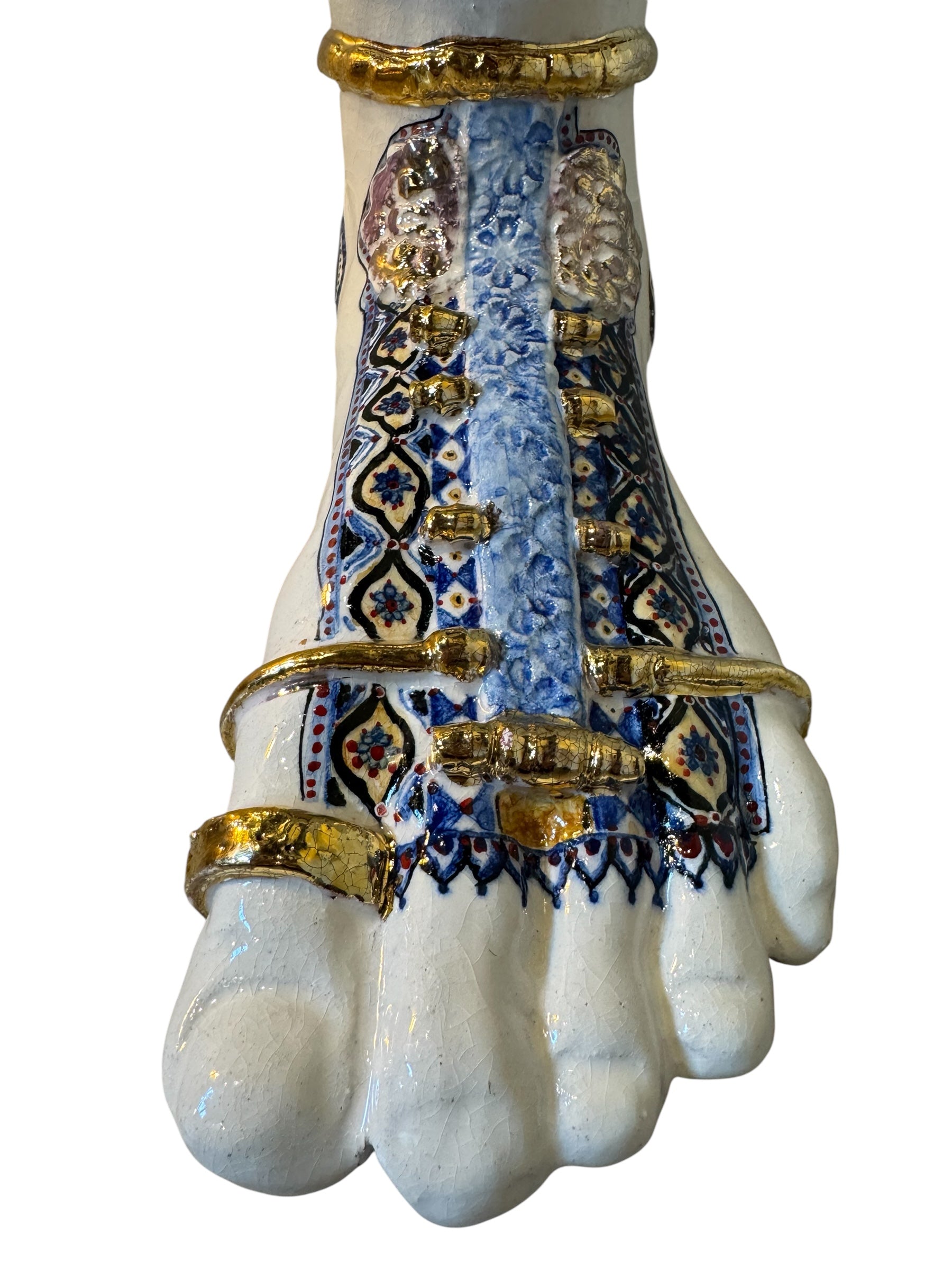Close-up of a ceramic foot sculpture by Domenico Pinto. The foot is decorated with intricate hand-painted designs inspired by Apulian folklore and accented with 24KT gold. A stylized gilt mask sits atop the foot.