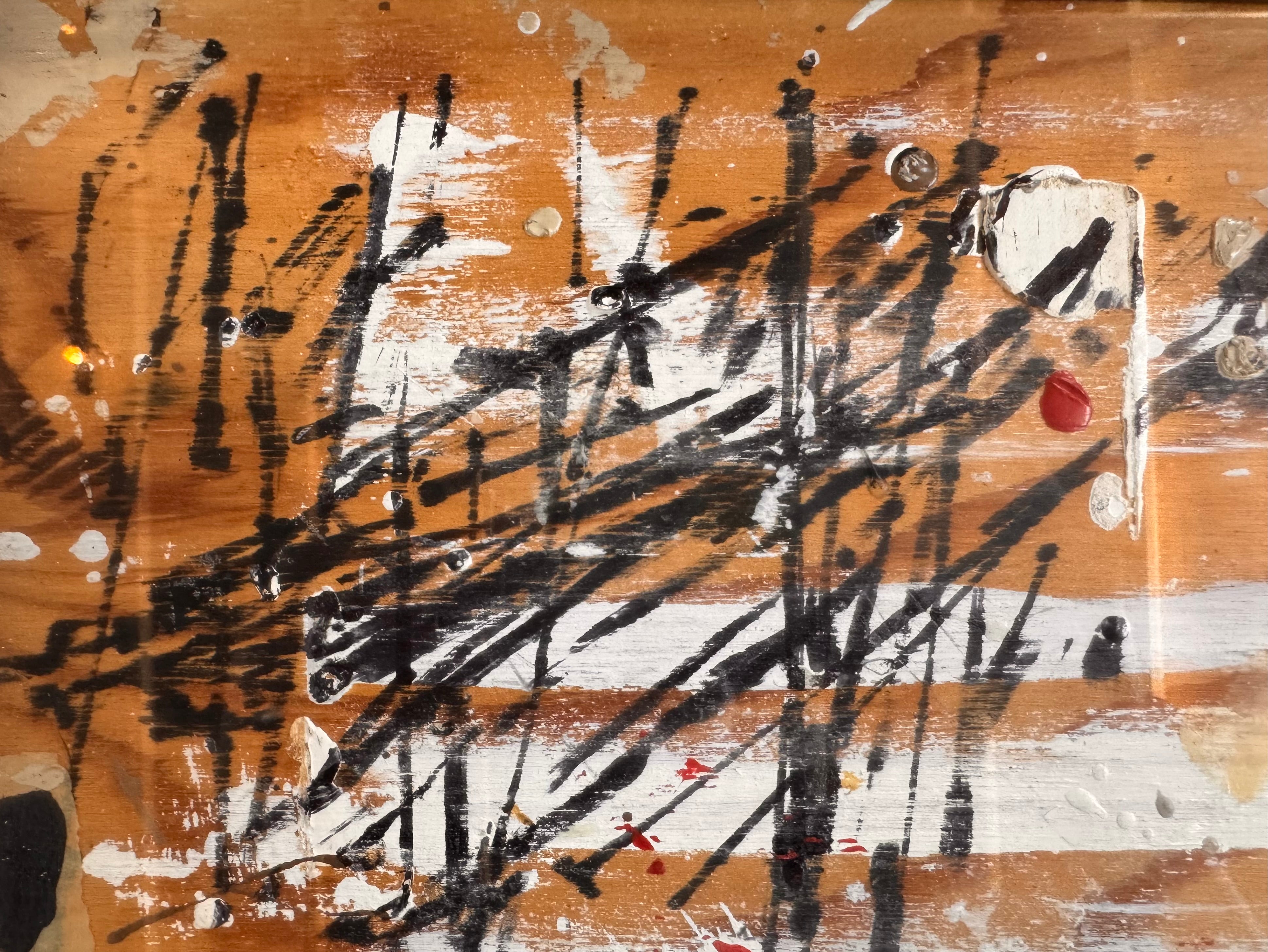 horizontal rectangular mixed media abstract artwork by J. Rogers, dated 2010, presented in a thin black frame. The composition is set against a light-toned wood grain background and features a dynamic, gestural arrangement of thick white impasto, splatters of red and black paint, and aggressive black scribbles. A vertical strip of textured material is collaged near the center, while various washes of tan and cream create a sense of weathered depth across the wooden surface.