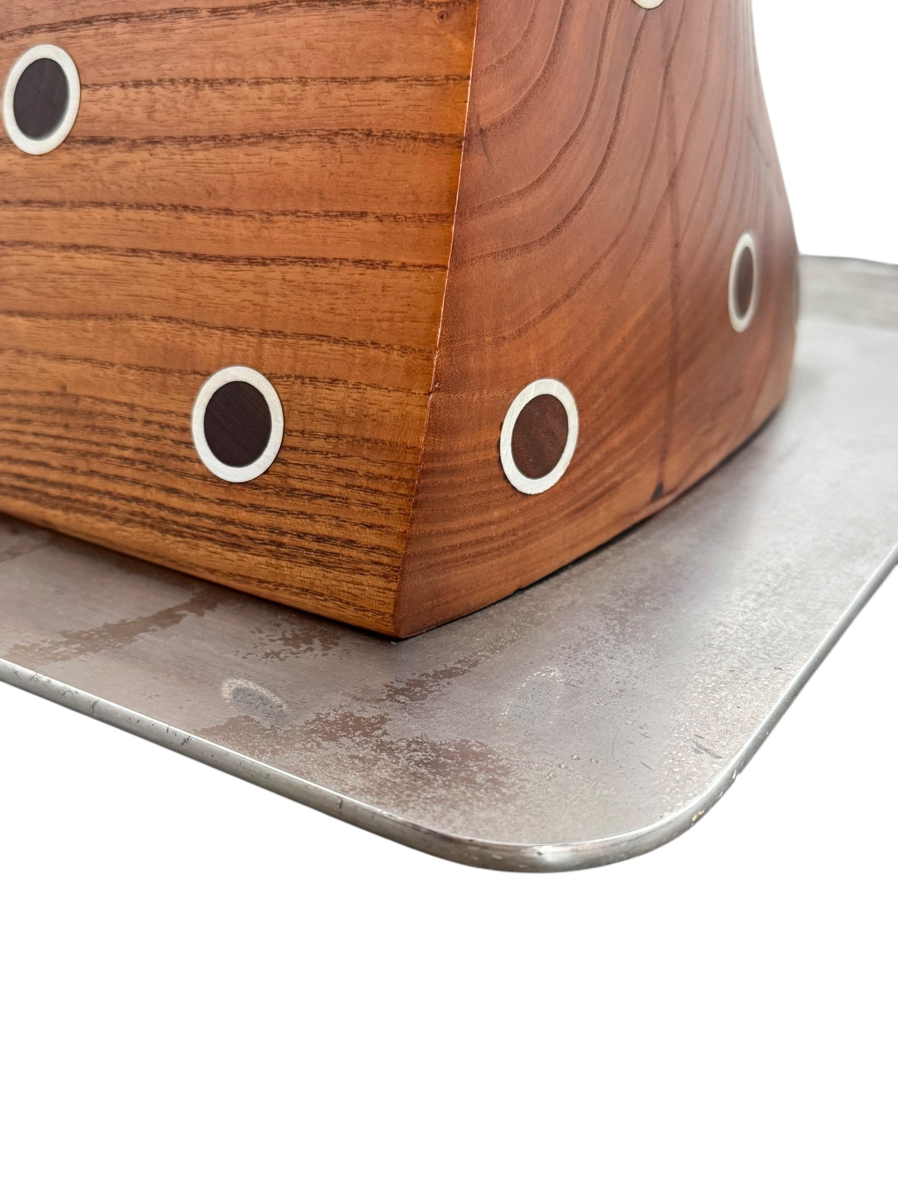 A wooden sculpture with a highly polished, reddish-brown grain, featuring two main asymmetrical, curved sections that converge at the top. Circular metal inlays with black centers are embedded across the surface of both wooden sections. A flat, polished metal blade-like element is inserted vertically between the two wooden sections, extending above the top surface. The sculpture rests on a metallic base.