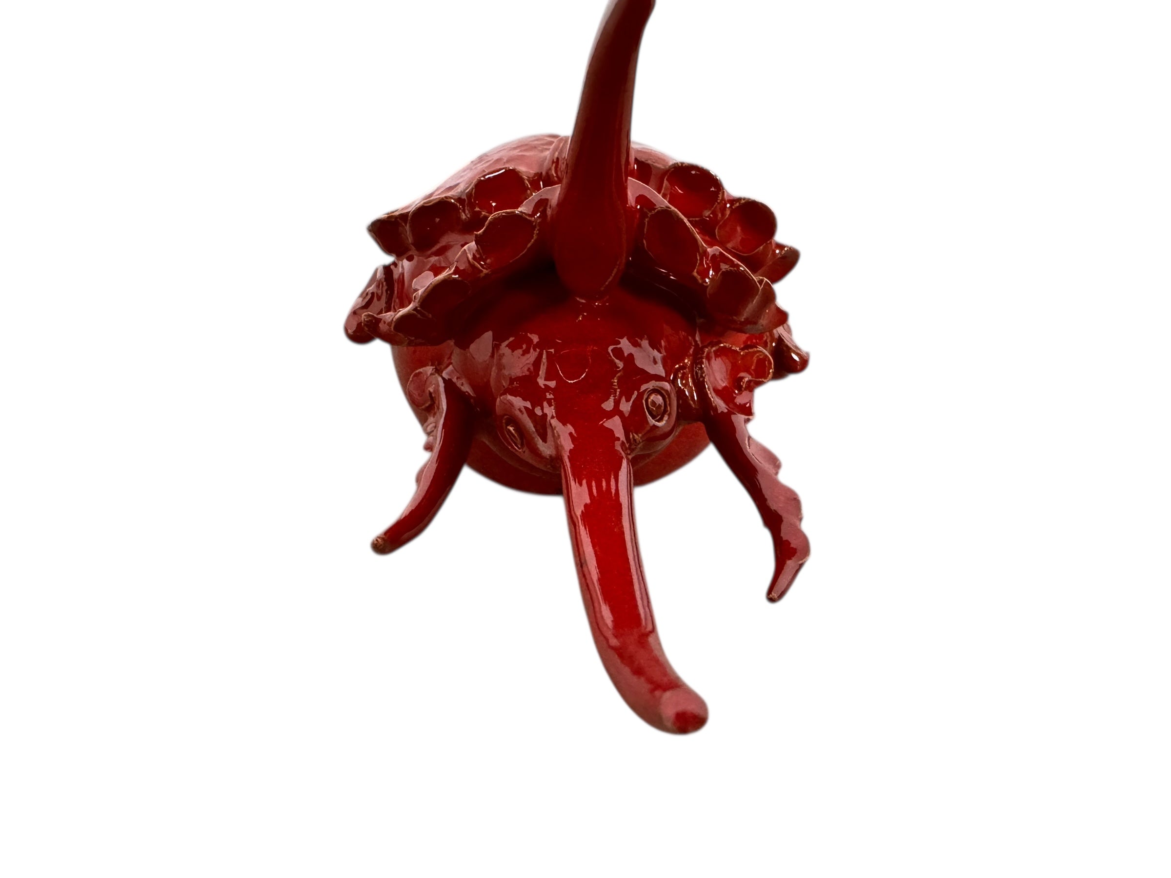 A handcrafted ceramic sculpture of a rhinoceros beetle by Enza Fasano, featuring a vibrant, glossy deep-red glaze. The piece is stylized with smooth, rounded contours and prominent, elegant horns extending forward. Its surface reflects light, highlighting the rich texture and artisanal detail of the clay.