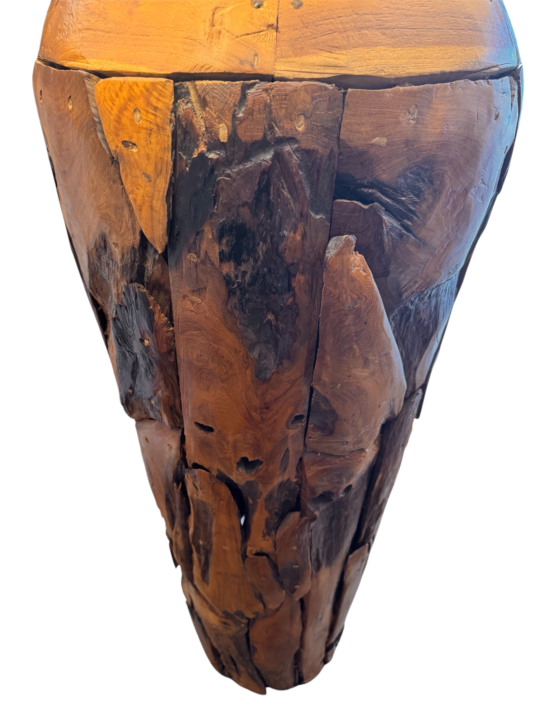 Two table lamps made from repurposed teak wood vases. The bases are tall and tapered, featuring a rugged, mosaic-like texture composed of various pieces of dark, polished teak wood with visible natural grain and crevices. Each lamp is topped with a large, warm-toned conical shade that is currently wrapped in protective clear plastic, emitting a soft, golden glow. The lamps are positioned on a clean white surface against a neutral background.