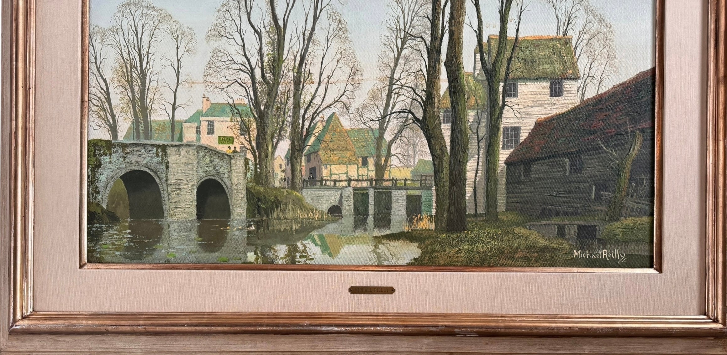 A naturalistic landscape oil painting by Michael Reilly titled "Mill Pond with Bridge" depicts a small town scene with a stone bridge over a mill pond, several buildings, and numerous bare trees under a grey sky