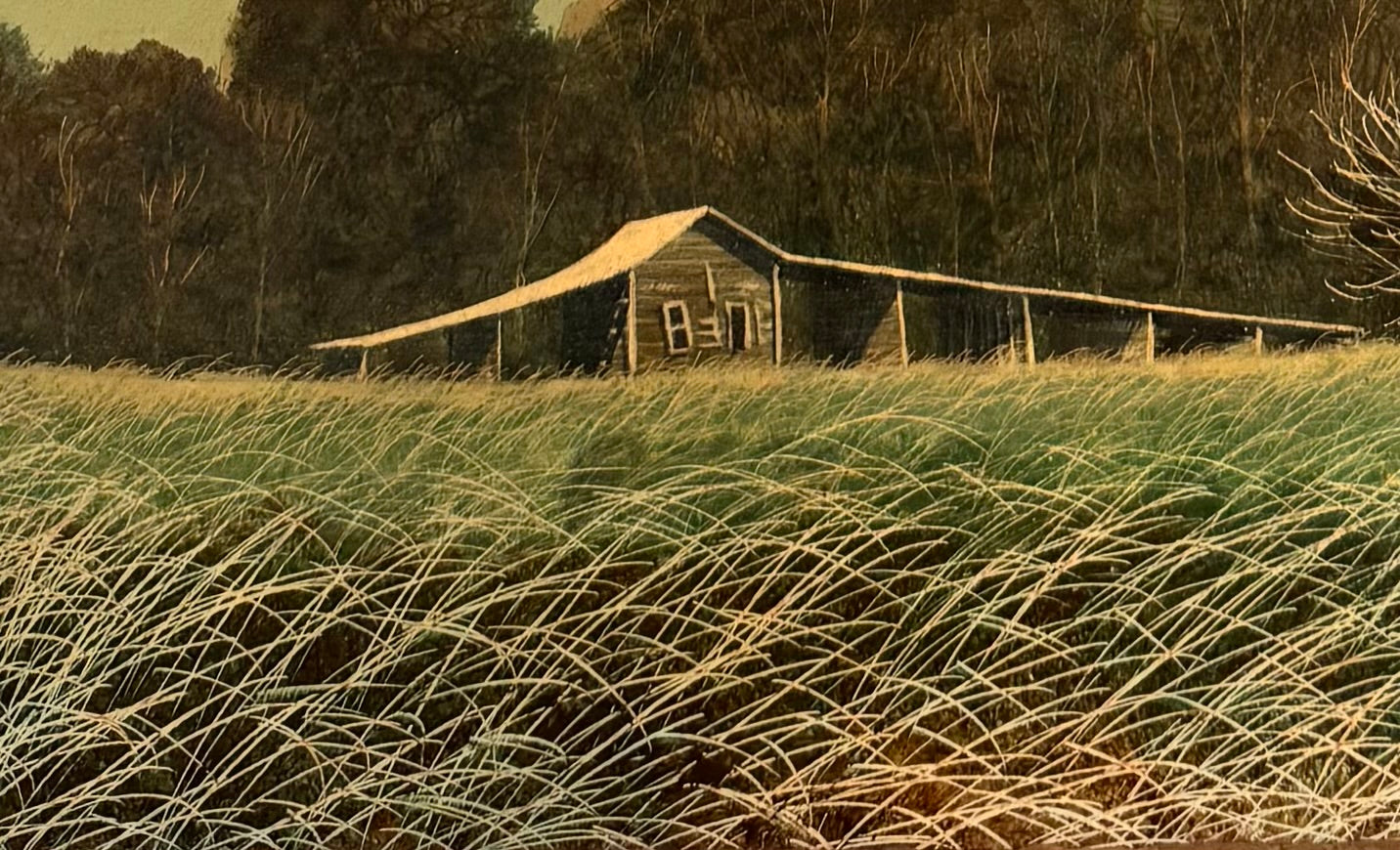 An oil painting on a wooden panel depicts a solitary scarecrow standing in a vast, grassy field under a pale yellow and blue sky. The scarecrow is shown from a three-quarters back view, wearing a brimmed hat and a tattered, patched brown coat with frayed straw bursting from its sleeves and edges. In the far left background, a long, white rustic barn sits before a dense line of green trees. The entire piece is enclosed in a thick, weathered, natural wood frame.