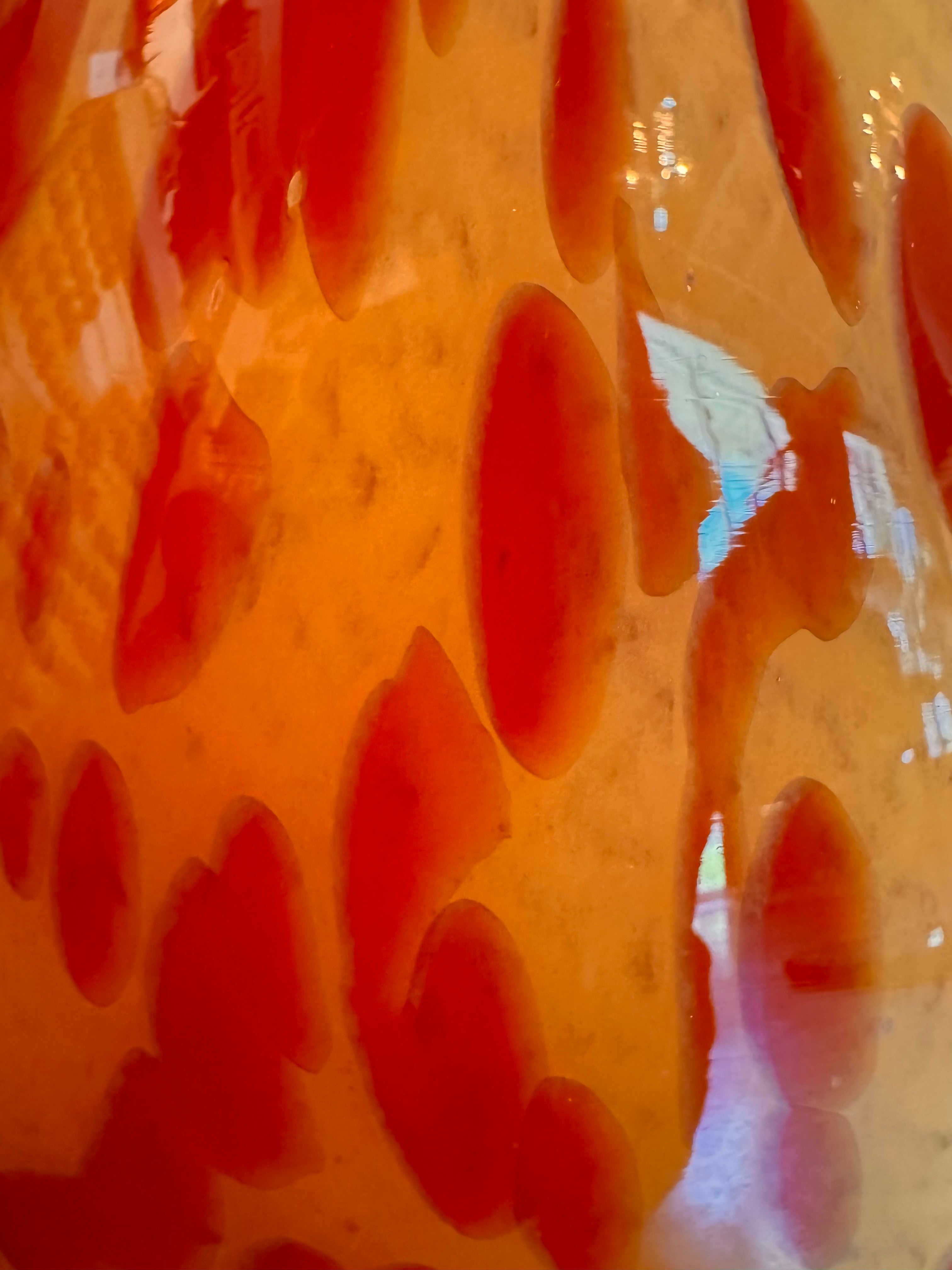 A hand-blown Italian Murano glass vase featuring a wide, bulbous body and a short, flared rim. The glass is a vibrant, translucent amber-orange base decorated with an organic "macchia" or spotted pattern of deep ruby red blotches. The red spots are elongated and vertical near the neck, becoming larger and more rounded toward the base. The surface has a high-gloss, reflective finish, showcasing mid-century modern craftsmanship.