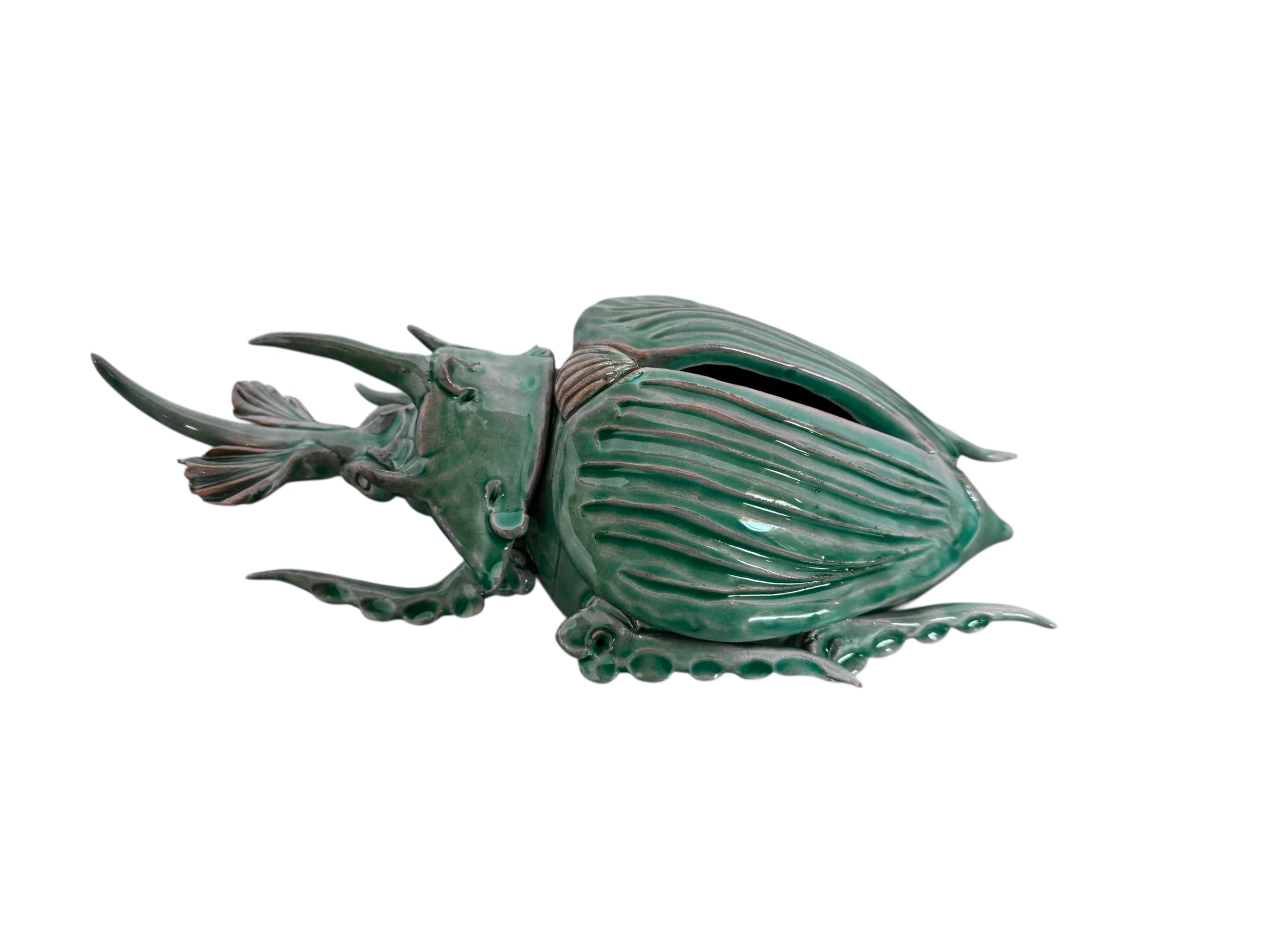 A ceramic sculpture of a stylized beetle by Enza Fasano, featuring a deep emerald green glaze with dark, metallic bronze-colored accents. The beetle is depicted with a highly textured, ribbed shell (elytra) and prominent, curved horns. Its legs are stylized with suckers, reminiscent of octopus tentacles. The piece has a glossy finish and rests against a plain white background.