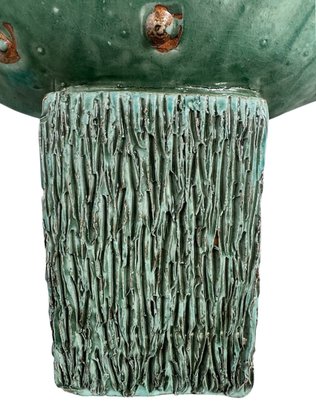 curved ceramic piece resembling a green pod, supported by a block plinth. The seafoam-green ceramic structure extends wide on either side, curving upward to form a wavy, boat-like shape. The outer surface is dotted with rows of small, raised, brown-orange, acorn-like protrusions. The top rim is layered with parallel, grooved ridges. The base is a vertical rectangular block with a heavily textured, bark-like surface in a dark green and black finish.