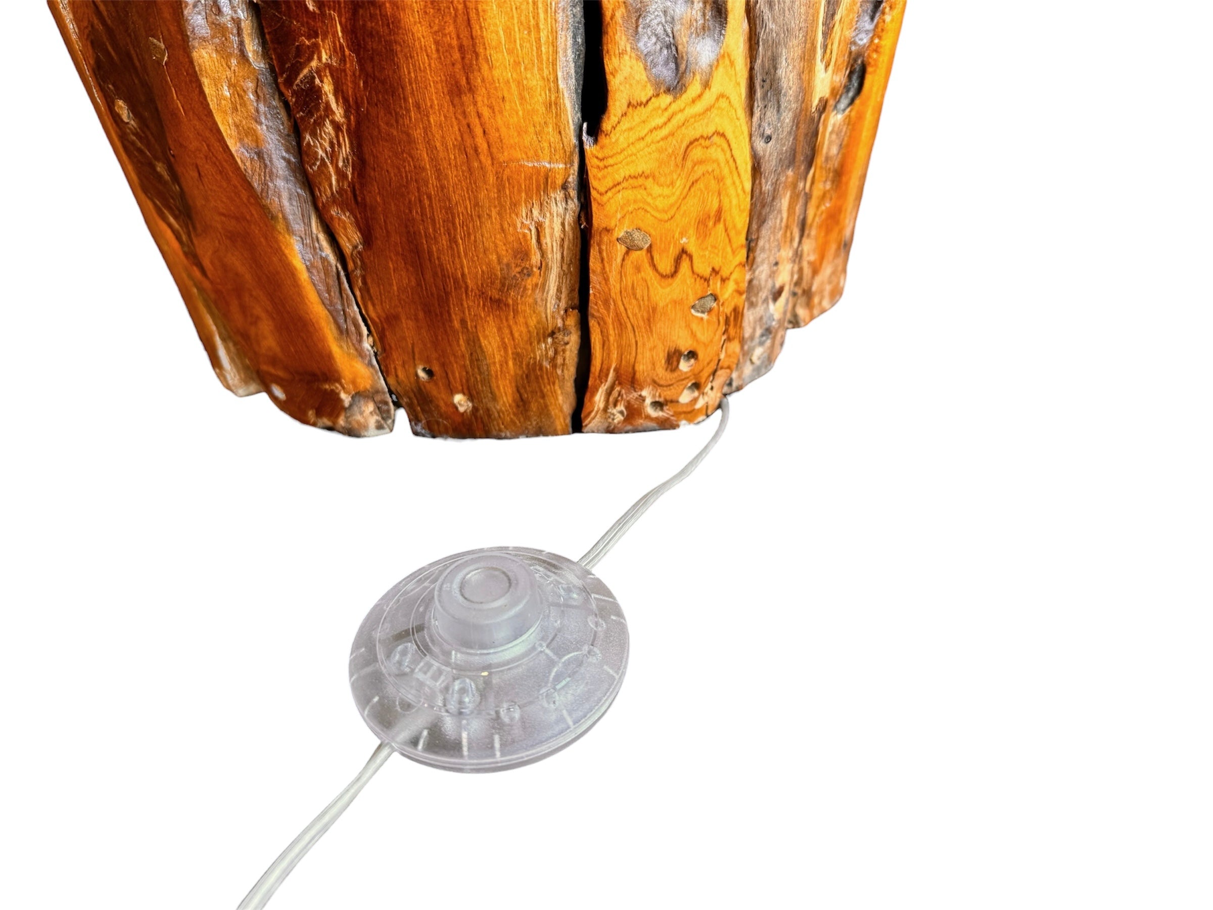 Two table lamps made from repurposed teak wood vases. The bases are tall and tapered, featuring a rugged, mosaic-like texture composed of various pieces of dark, polished teak wood with visible natural grain and crevices. Each lamp is topped with a large, warm-toned conical shade that is currently wrapped in protective clear plastic, emitting a soft, golden glow. The lamps are positioned on a clean white surface against a neutral background.