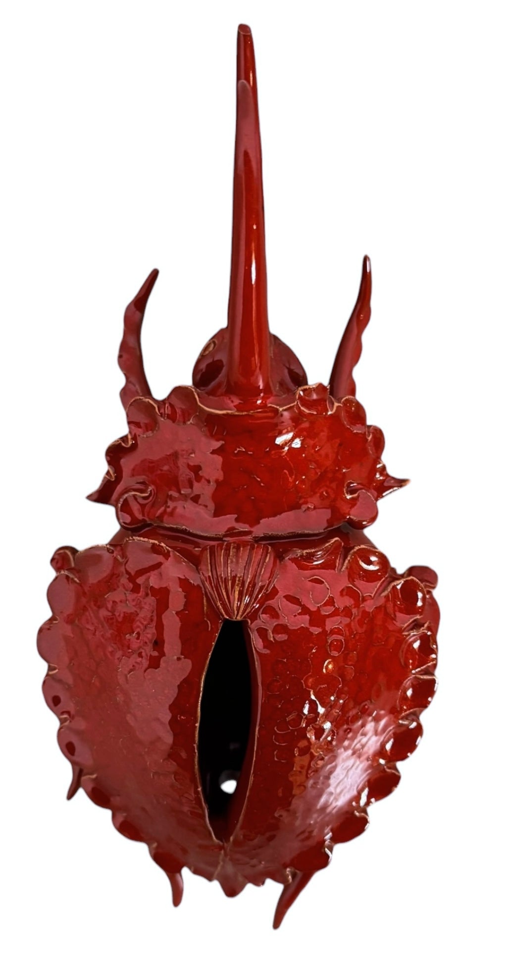 A handcrafted ceramic sculpture of a rhinoceros beetle by Enza Fasano, featuring a vibrant, glossy deep-red glaze. The piece is stylized with smooth, rounded contours and prominent, elegant horns extending forward. Its surface reflects light, highlighting the rich texture and artisanal detail of the clay.