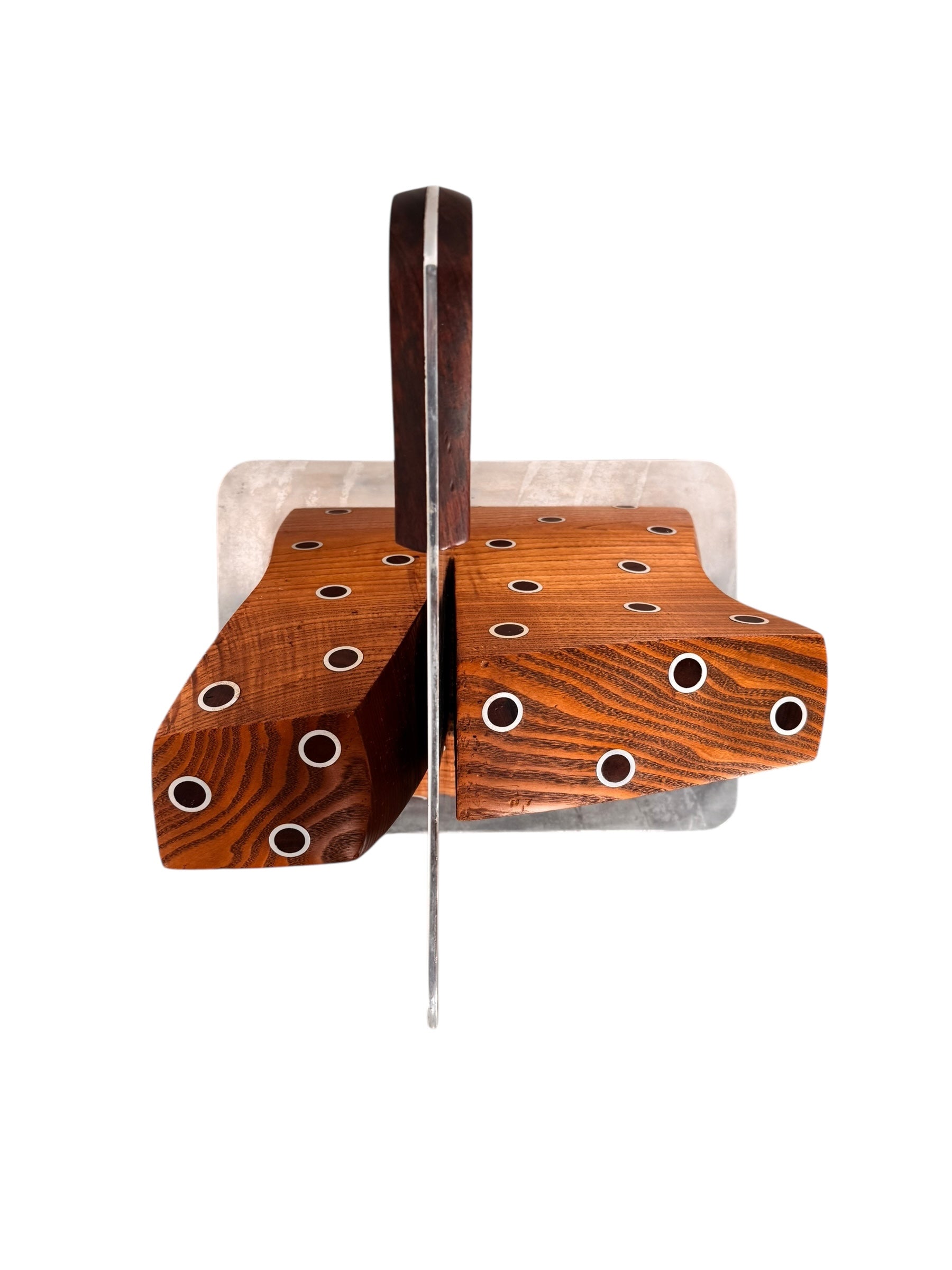 A wooden sculpture with a highly polished, reddish-brown grain, featuring two main asymmetrical, curved sections that converge at the top. Circular metal inlays with black centers are embedded across the surface of both wooden sections. A flat, polished metal blade-like element is inserted vertically between the two wooden sections, extending above the top surface. The sculpture rests on a metallic base.