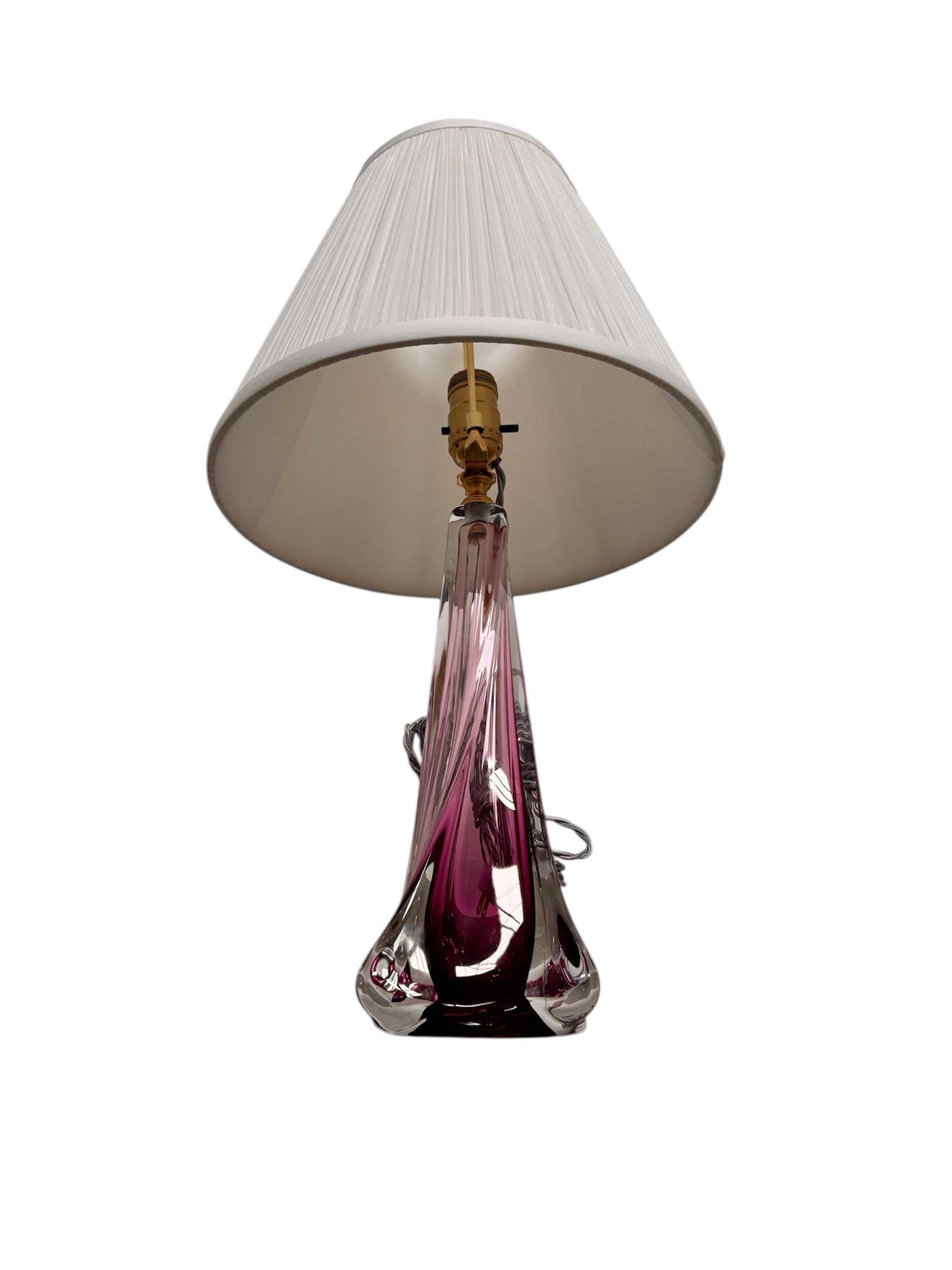 A tall, Mid-Century Modern sculptural table lamp featuring a hand-blown art glass base. The base is crafted in the Sommerso style, with a deep amethyst purple core encased in a thick, clear crystal outer layer that twists upward in an organic, fluid motion. The lamp is topped with a white, knife-pleated empire shade and a clear glass ball finial. The overall design is elegant and slender, emphasizing the contrast between the saturated purple glass and the transparent, light-refracting crystal.