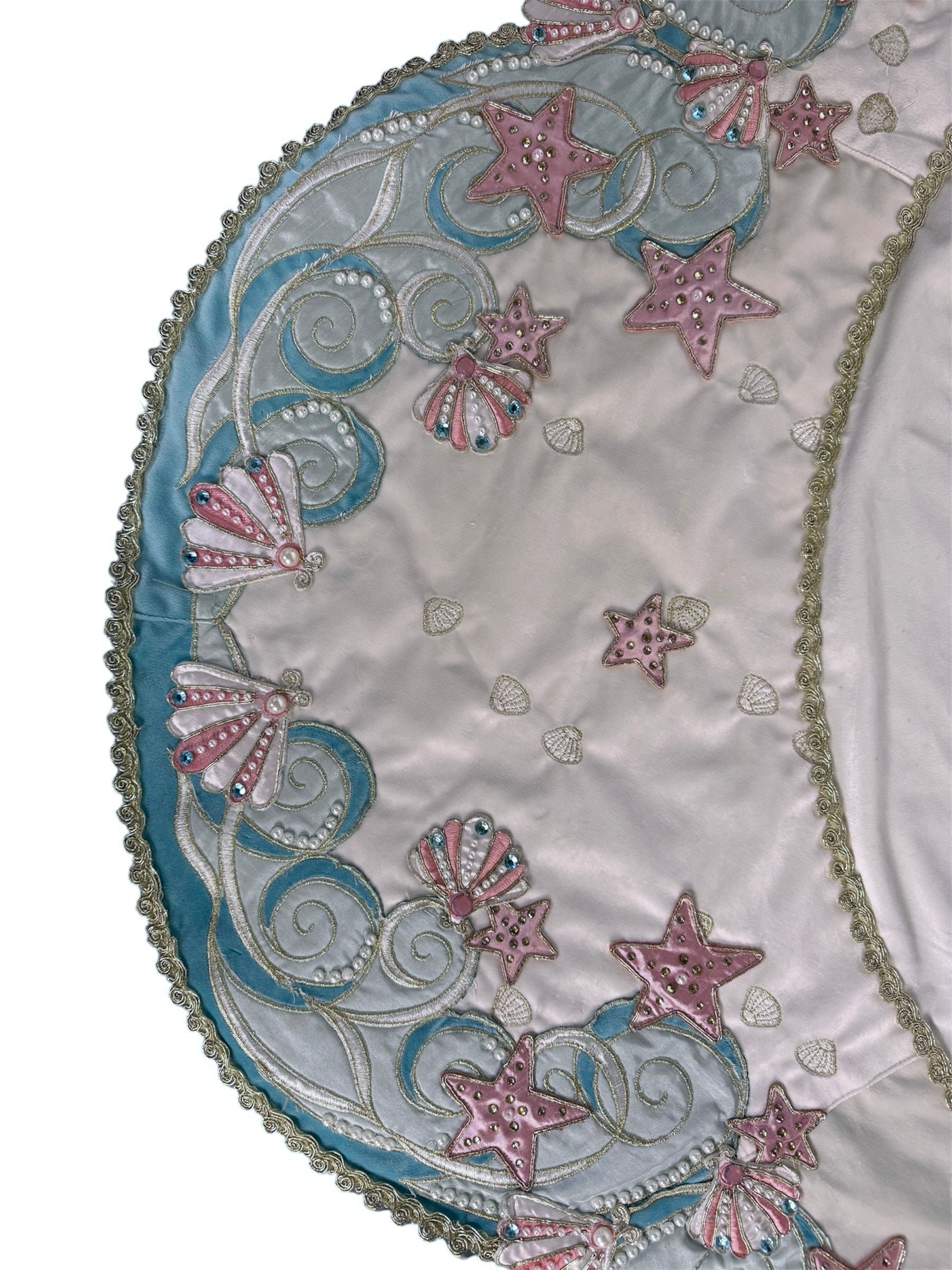 A round, cream-colored tablecloth featuring a dramatic, scalloped border shaped like six large flower petals. The border is intricately embroidered with a coastal motif including pink starfish, seashells, and teal-blue swirling waves. Gold beaded accents line the inner circular seam and the outer edges.
A long, narrow cream runner with matching scalloped, wavy edges. It is adorned with the same nautical embroidery of pink starfish and seashells scattered across the center and concentrated along the edge.