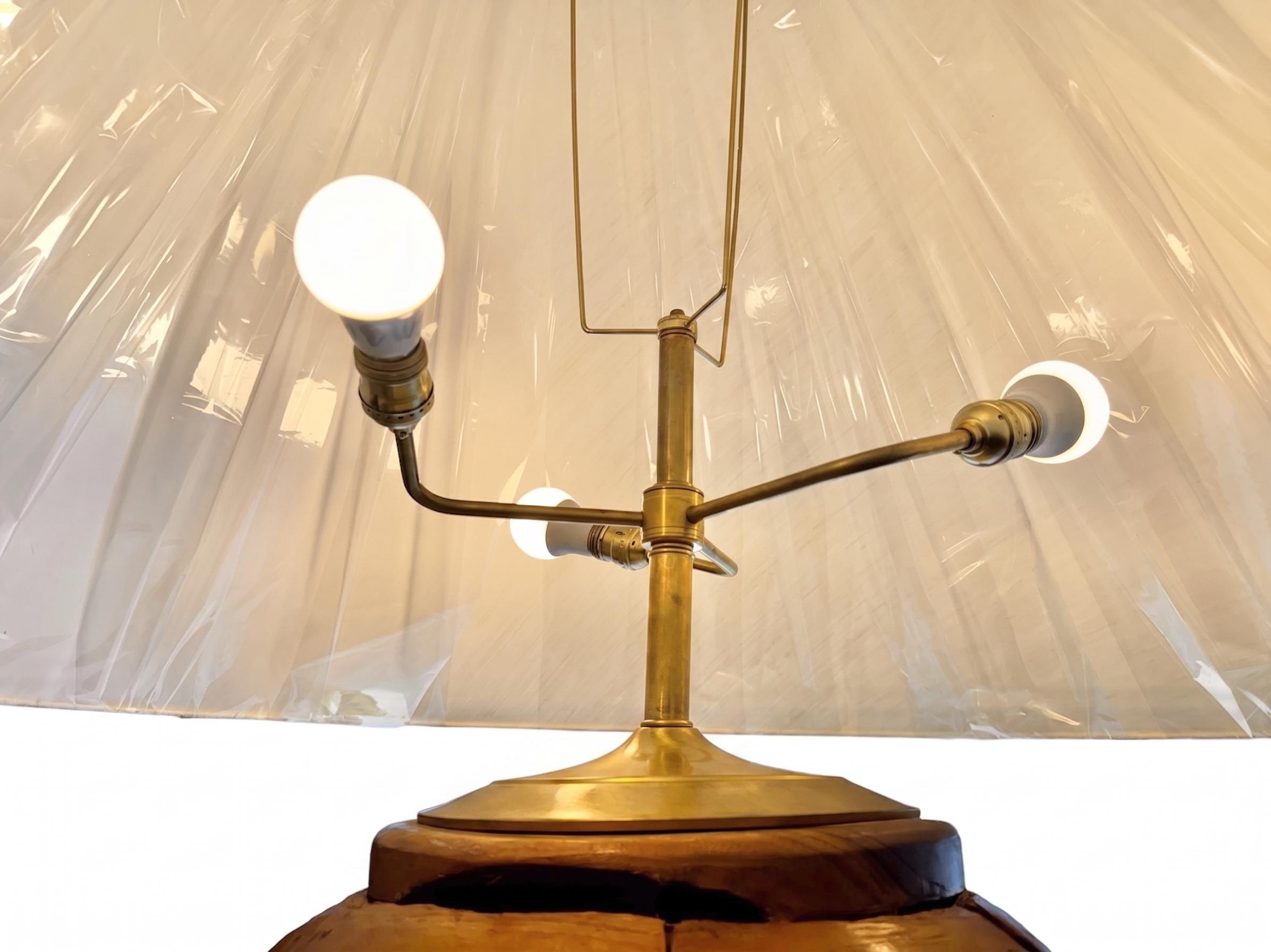 Two table lamps made from repurposed teak wood vases. The bases are tall and tapered, featuring a rugged, mosaic-like texture composed of various pieces of dark, polished teak wood with visible natural grain and crevices. Each lamp is topped with a large, warm-toned conical shade that is currently wrapped in protective clear plastic, emitting a soft, golden glow. The lamps are positioned on a clean white surface against a neutral background.