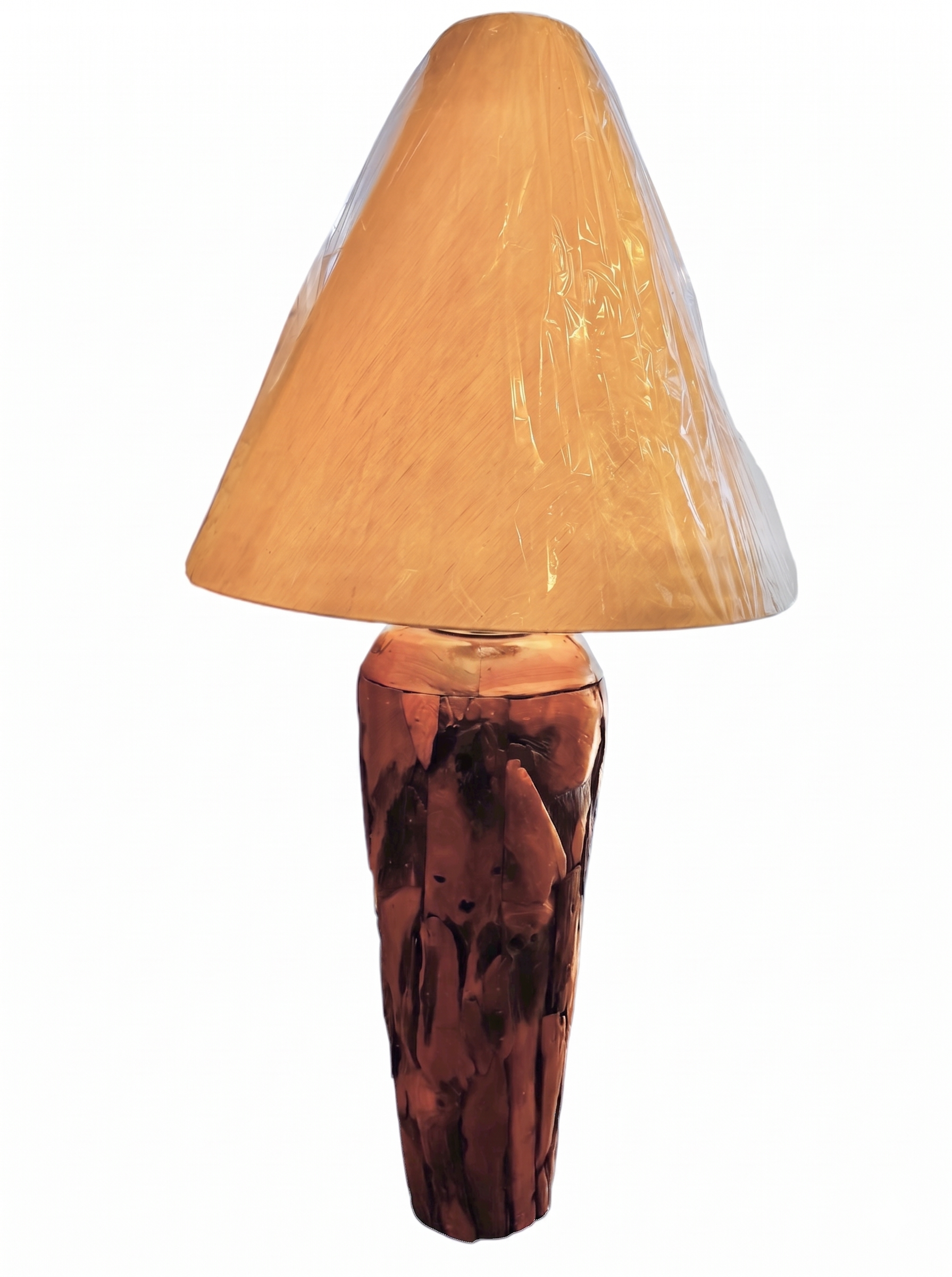 Two table lamps made from repurposed teak wood vases. The bases are tall and tapered, featuring a rugged, mosaic-like texture composed of various pieces of dark, polished teak wood with visible natural grain and crevices. Each lamp is topped with a large, warm-toned conical shade that is currently wrapped in protective clear plastic, emitting a soft, golden glow. The lamps are positioned on a clean white surface against a neutral background.