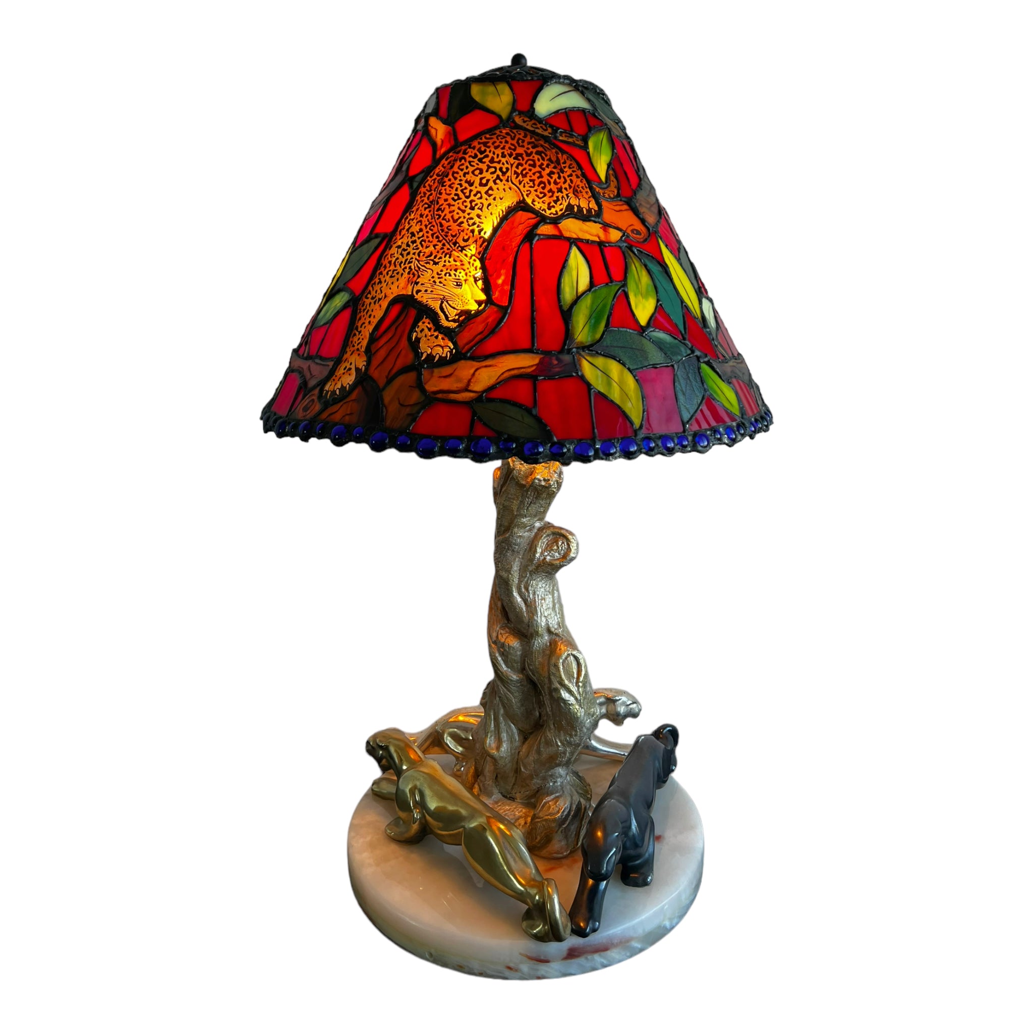 A lamp with a marble base featuring a sculpture of three jaguars and a bespoke stained glass shade with a depiction of cheetahs. The lampshade has a colorful design with a prominent red and yellow motif.