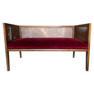 A mid-century cane settee with a burgundy velvet upholstered seat and a cane back and armrests in a walnut finish.